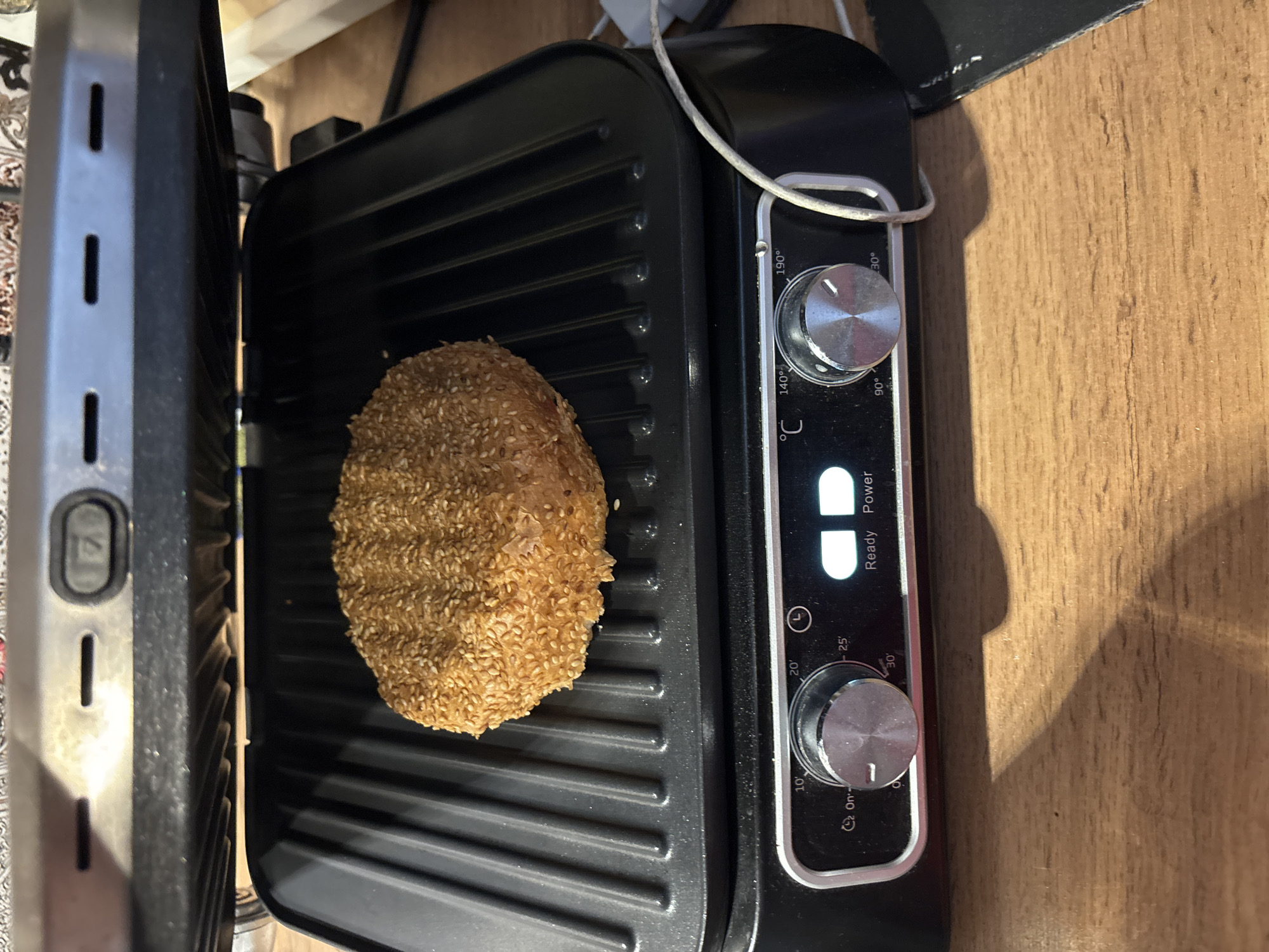 A sesame seed bagel is being grilled on an open electric grill. The grill is black with silver knobs and a digital display. The bagel is round and appears to be golden brown and slightly toasted. The grill is placed on a wooden surface. The image does not provide any information about the location in Kemerovo, Russia.