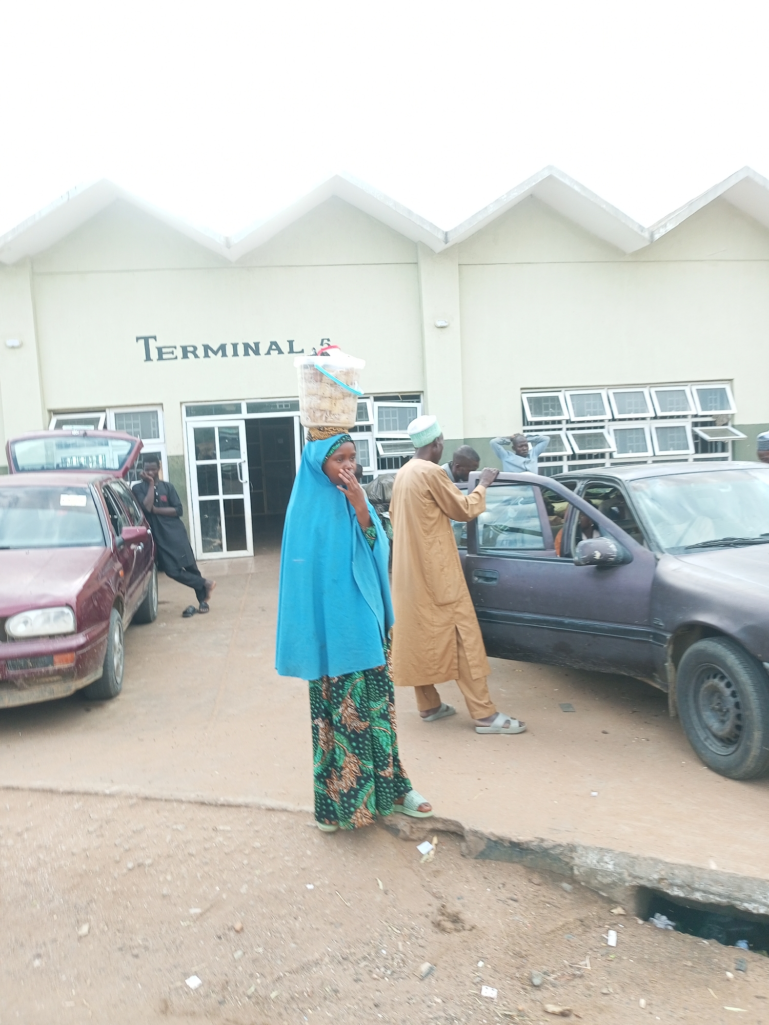 The image depicts the entrance to "TERMINAL 6" in Gombe, Nigeria. A woman wearing a blue hijab and a patterned dress, carrying a large plastic container of food items on her head, stands in the foreground, looking toward the viewer and touching her face. To her left, a maroon car with its trunk open is parked, and a young man in dark clothing stands leaning against it. To the right of the woman, a dark-colored sedan is parked, with its driver's side door open. A man in a light brown robe and a white cap stands by the open car door, looking inside. Behind him, another man in a light blue shirt has his hands behind his head. The building behind them has a segmented roofline and numerous windows. The ground is dusty and appears to be a road or parking area.