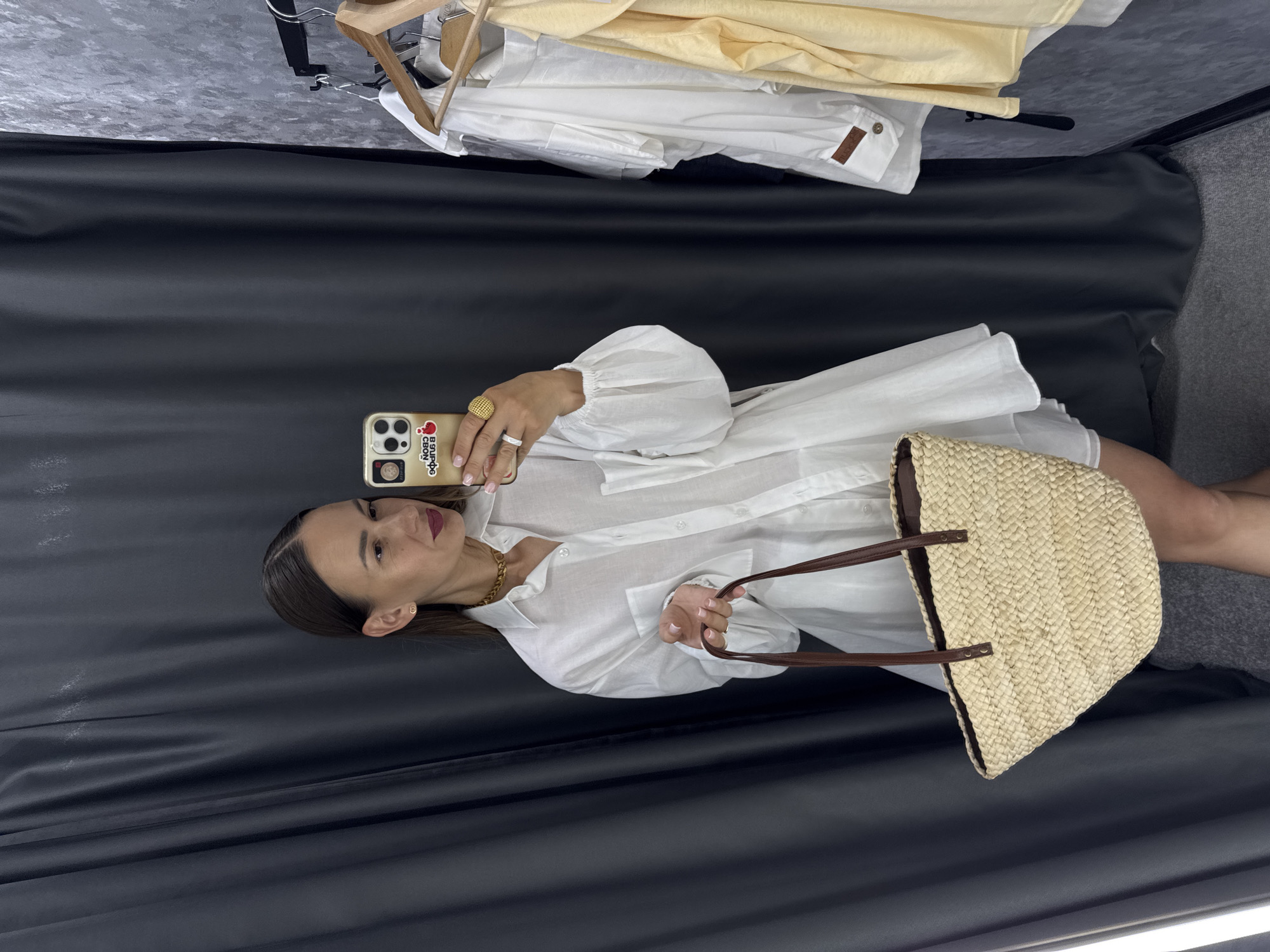 A young woman stands in a dressing room, taking a mirror selfie. She is wearing a white oversized shirt, a gold chain necklace, and a large gold ring on her right hand. She holds a straw bag with brown leather straps in her left hand. In the background, to the right, several garments are hanging on a rack, including white shirts and a yellow top. The dressing room features dark grey curtains and a grey textured wall. The image appears to be taken indoors, possibly in a retail store or a home dressing room. The location is identified as Sochi, Russia.