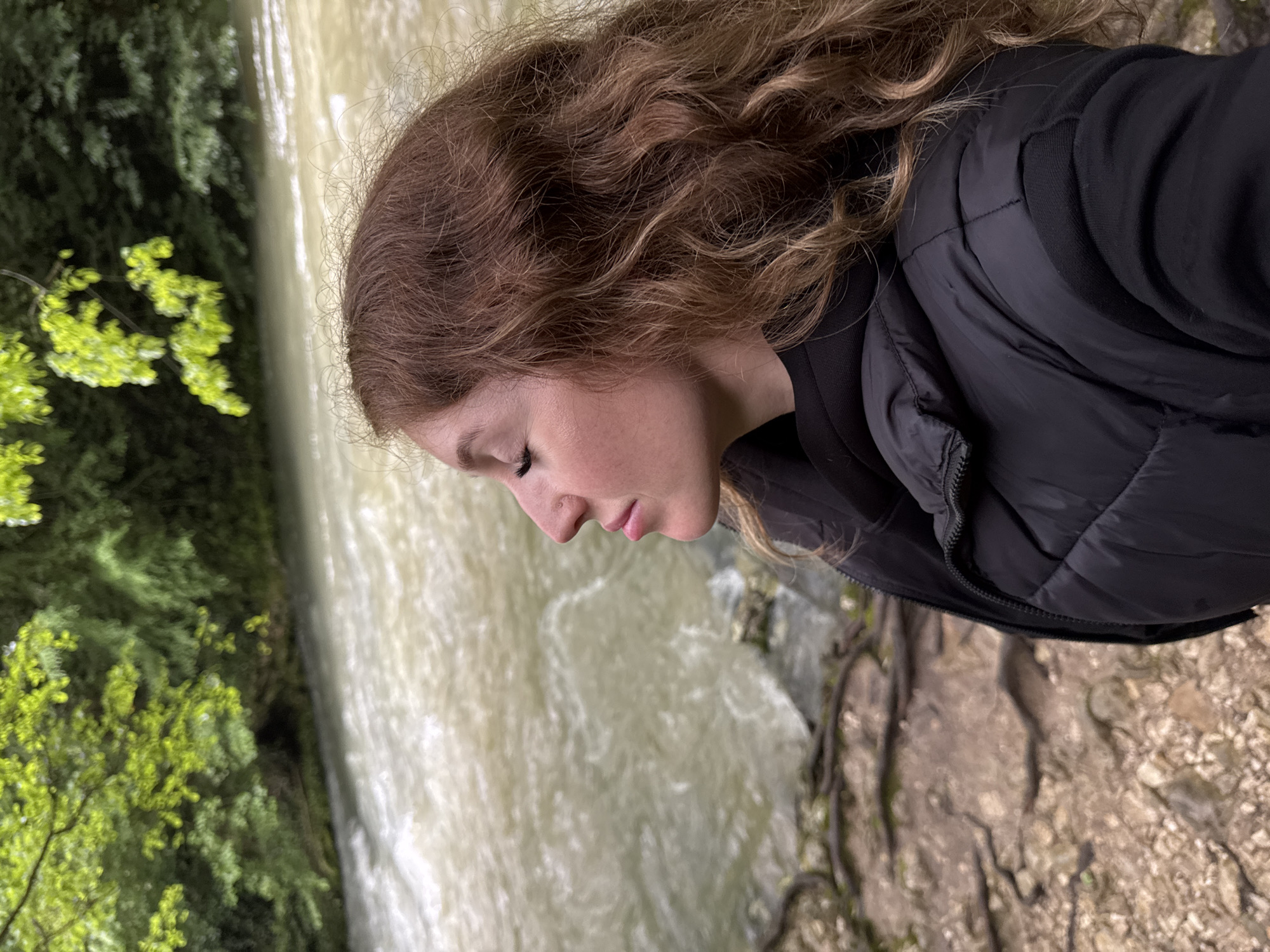 The image depicts a woman with brown curly hair, wearing a black jacket, standing outdoors. She is looking down, seemingly contemplative. Behind her is a body of water, likely a river or stream, with turbulent, light-colored water. Green foliage is visible on the left side of the frame, suggesting a natural environment, possibly near Tsabal, Abkhazia. The right side of the image reveals what looks like rocky terrain with exposed roots.