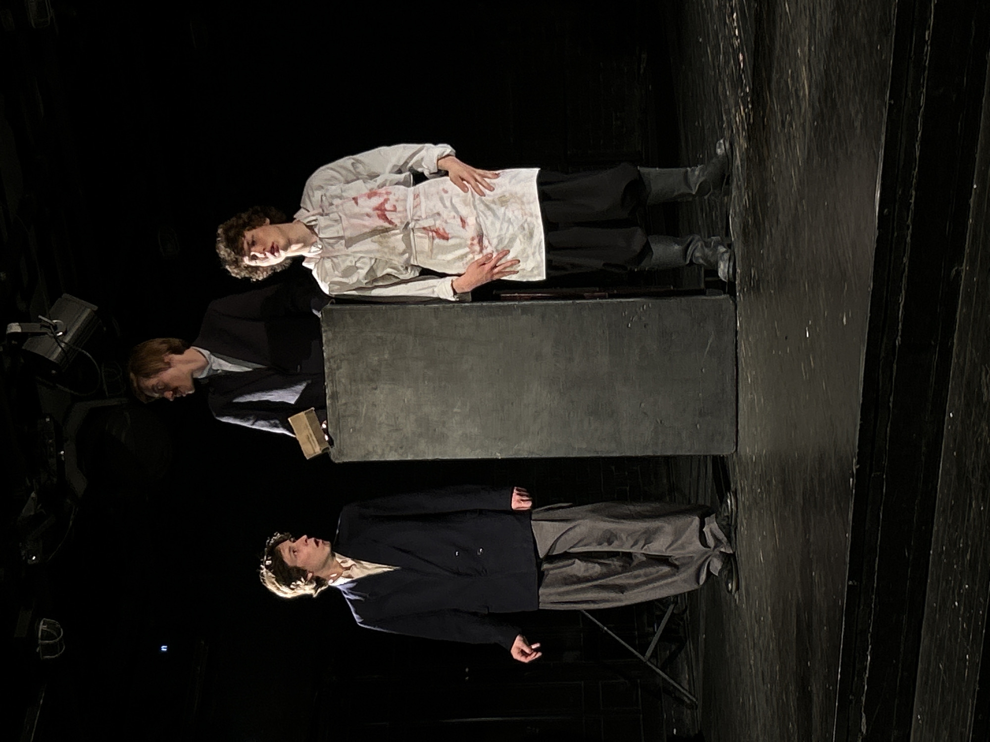 This image captures a theatrical scene on a dark stage in the city of Unknown, Unknown. Three young males are present, illuminated against a dark background, suggesting artificial stage lighting.
On the left, a male with light brown, wavy hair wears a light-colored collared shirt, a dark navy double-breasted blazer, and loose-fitting grey trousers with dark shoes. His mouth is slightly open, and his gaze is directed to the right, appearing surprised or in reaction. His right hand is slightly raised with fingers splayed.
In the center, partially obscured by a dark, rectangular podium-like structure, stands another male with light brown hair and a faint mustache. He wears a light-colored collared shirt and a dark suit jacket. His left hand rests on the podium, holding a small, light brown, rectangular object, possibly a book or box. His gaze is directed downwards and to the right, away from the camera.
On the right, a third male with curly dark brown hair wears a white long-sleeved shirt and a white apron. The apron and parts of the shirt are heavily stained with red splatters. He wears dark, loose-fitting trousers tucked into dark, knee-high boots. His hands rest at his sides, and his mouth is slightly open with his gaze directed upwards and to the left.
The stage floor is dark, possibly wooden, with subtle texture visible. Overhead, stage lighting equipment is partially discernible against the black background. A dark metal support structure is partially visible on the far left of the stage. The individuals are positioned to create a dramatic triangle, indicating an ongoing performance or scene.