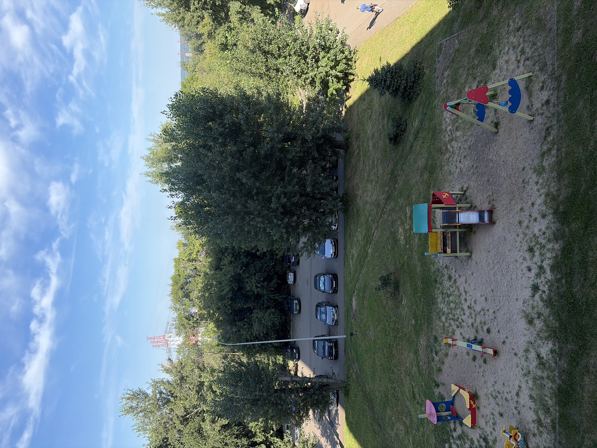The image captures a view of a playground and parking lot in Naberezhnye Chelny, Russia. In the foreground, a sand-covered playground features a red and blue swing set, a yellow and red multi-part play structure with a slide, a seesaw, and a sandbox with a small yellow toy vehicle. Beyond the playground, a grassy area slopes upwards towards a paved parking lot filled with parked cars. Trees surround the parking lot and extend into the background. In the far distance, a large red and white power pylon is visible against a blue sky with scattered clouds. A single individual in a blue shirt and shorts walks on a paved path to the right of the image. The scene appears to be a typical residential area.