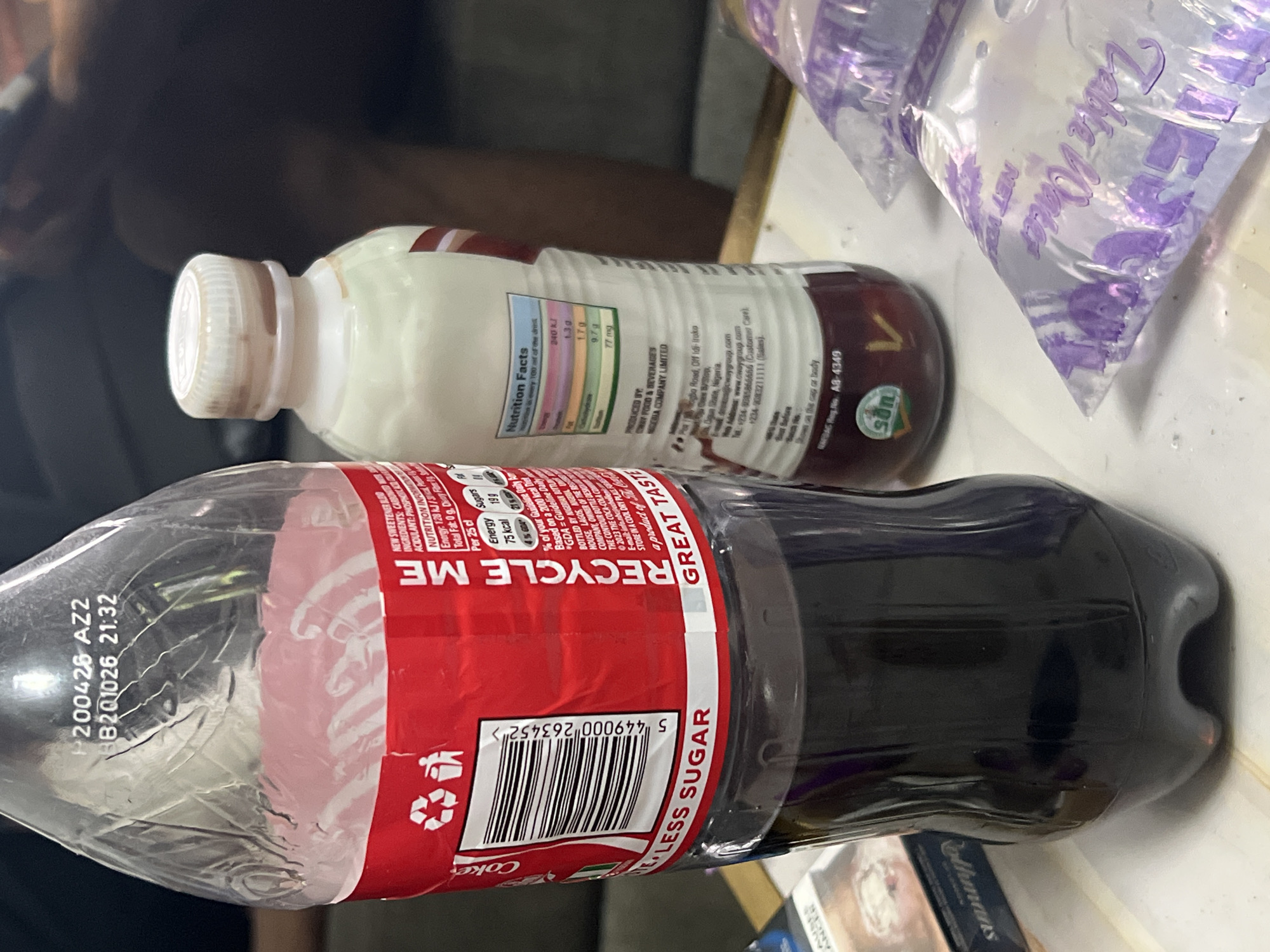 The image shows a tabletop with several items on it. In the foreground, there is a plastic Coca-Cola bottle labeled "LESS SUGAR" with a red and white label and barcode, partially filled with a dark liquid. Behind the Coca-Cola bottle is a smaller, white plastic bottle with a white cap; the label contains "Nutrition Facts" and production information, including "CWAY FOOD & BEVERAGES MERIA COMPANY LIMITED". Behind these bottles is a clear plastic bag filled with water and printed with purple text that reads, "Table Water". Adjacent to the coke bottle is a packet of cigarettes. Based on the labels, the items suggest a location in Nigeria, specifically Abuja. It is a close-up image with shallow depth of field.