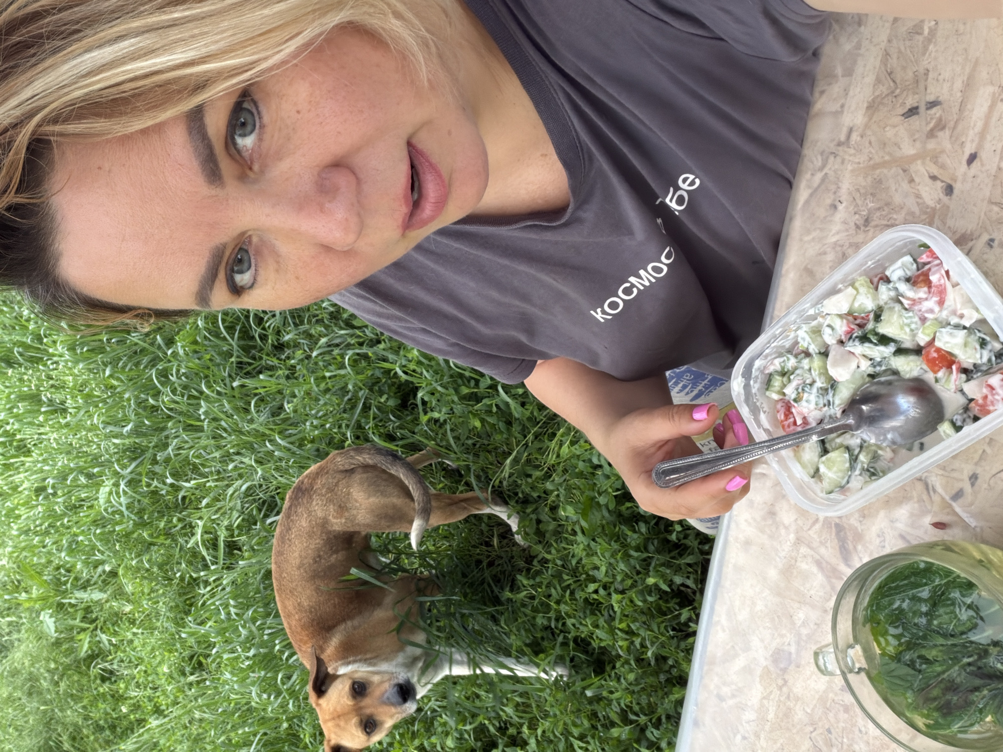 The image depicts a woman outdoors, possibly in Leningradskaya Oblast, Russia, given the likely transliteration of "Kocmoc" on her grey t-shirt, potentially suggesting a connection to space programs associated with Russia, often linked to the region. The woman is holding a container of salad with a spoon. A brown dog is standing in the grass nearby. The location appears to be on a lawn with tall grass. There is a clear glass jar filled with green leaves and liquid sitting on a wooden surface next to the salad. The woman is looking at the camera. Her fingernails are painted pink. The image seems to be a self-portrait or taken at close range by another person. The environment is natural.