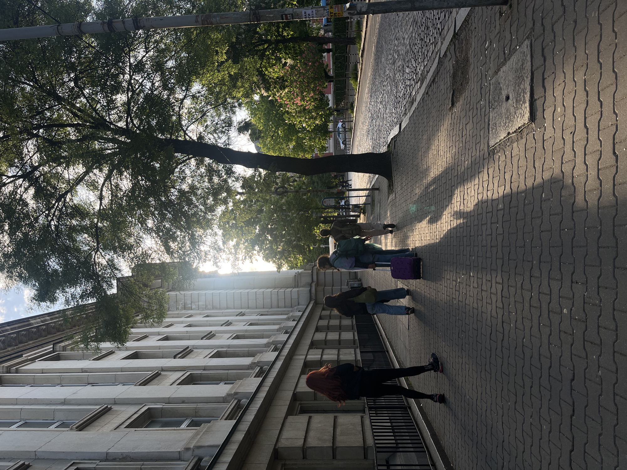 The image shows a street scene in Sofia, Bulgaria, with several pedestrians walking along a sidewalk next to a large building. The building is light-colored with multiple windows and architectural details. A tree with dense foliage extends over the sidewalk, casting shadows. The sidewalk is paved with dark gray interlocking blocks. People are walking, some with luggage. The lighting suggests it is either early morning or late afternoon, with the sun low in the sky. A cobbled street is visible on the other side of the sidewalk.