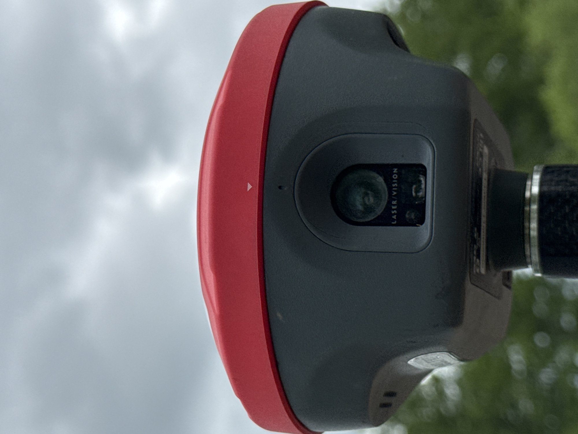 The image shows a close-up of a red and gray surveying device, possibly a total station or laser scanner, mounted on a pole. The device has a prominent red top and a dark gray body with a central lens assembly labeled "LASER/VISION." In the background, the sky is overcast with gray clouds, and blurred green foliage suggests an outdoor, possibly urban or suburban, environment. The device is positioned outdoors, and the lighting conditions indicate it is daytime. There are no people visible in the image. The location of Муртыгит, Russia is not depicted or suggested by any visual cues in the provided image.