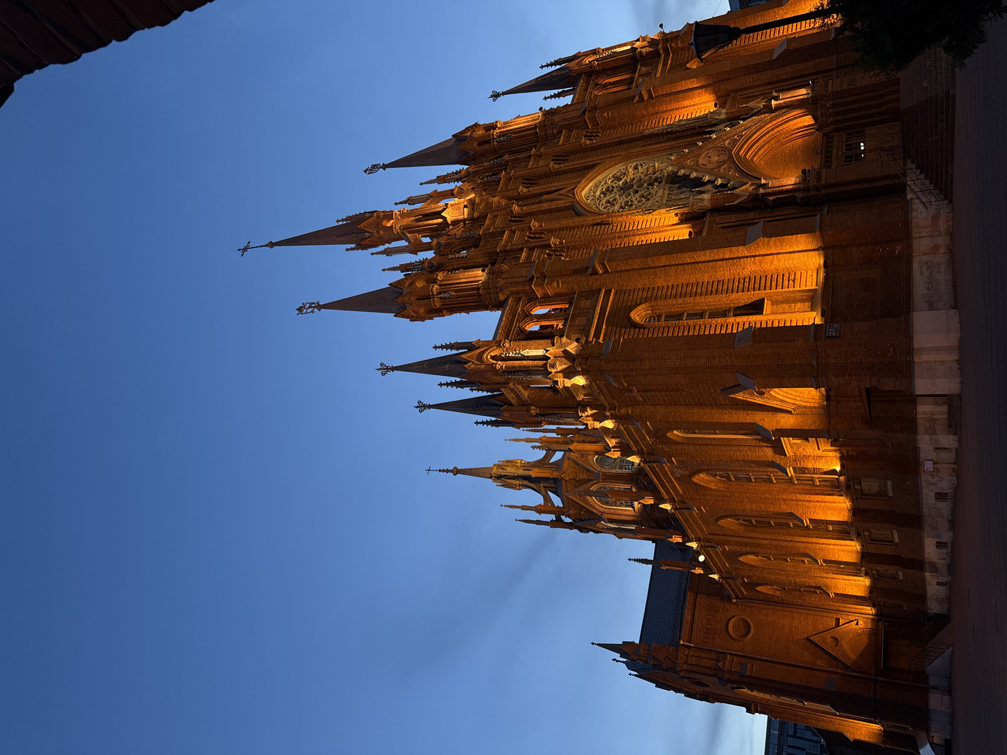 The image depicts a brick cathedral with multiple spires against a twilight sky, illuminated by warm artificial light. The architectural style is Gothic Revival. The cathedral appears to be located in Moscow, Russia, based on the architectural features and overall ambiance. There are no people visible in the frame. The environment is urban, given the presence of a street or sidewalk in the foreground. The scene is still and quiet, likely captured during evening hours.