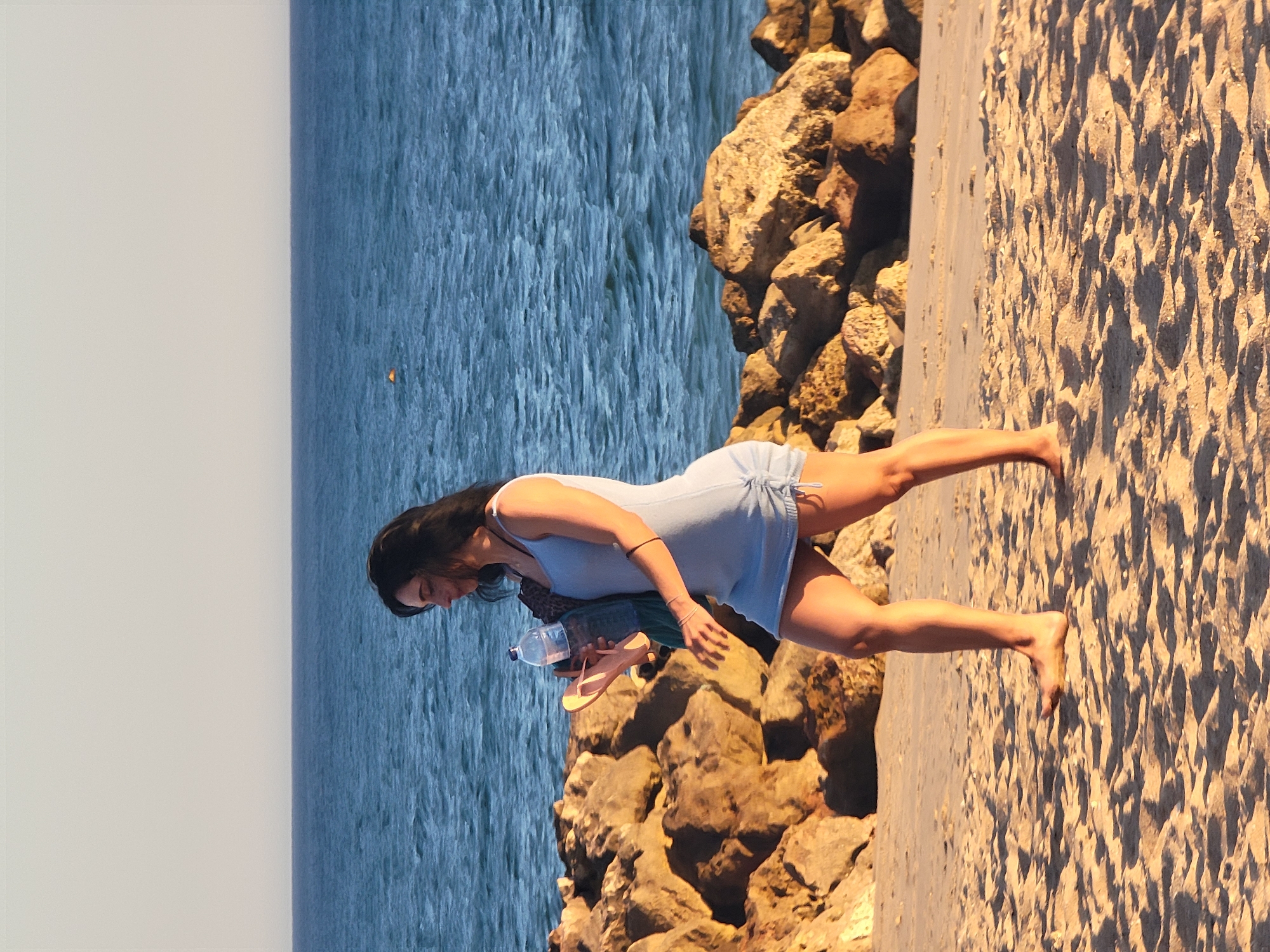 A woman walks along the sandy beach in Quarteira, Portugal, near a rocky breakwater. She is wearing a light blue tank dress and carrying a plastic water bottle and a pair of flip-flops. The ocean stretches out behind her, with a clear, pale sky overhead. The sun is casting long shadows on the sand, indicating it is either early morning or late afternoon.