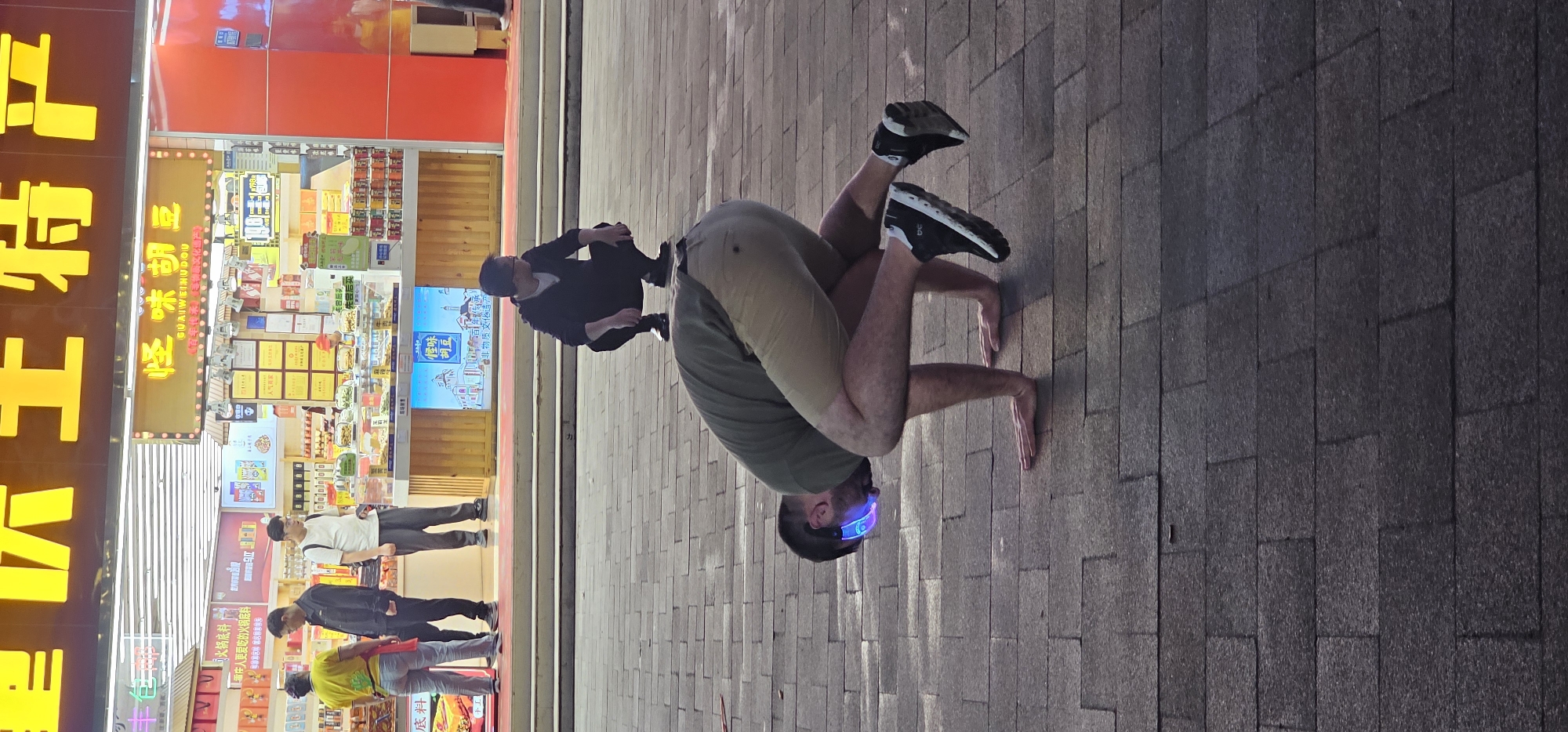 On a paved surface in Yuzhong District, China, a man wearing tan pants, an olive green t-shirt, and black and white running shoes is performing a handstand. He is wearing a blue LED device on his head. In the background, another man in a black jacket and mask is squatting. Further in the background, pedestrians walk past a store with a red and orange facade featuring yellow Chinese characters and product displays.
