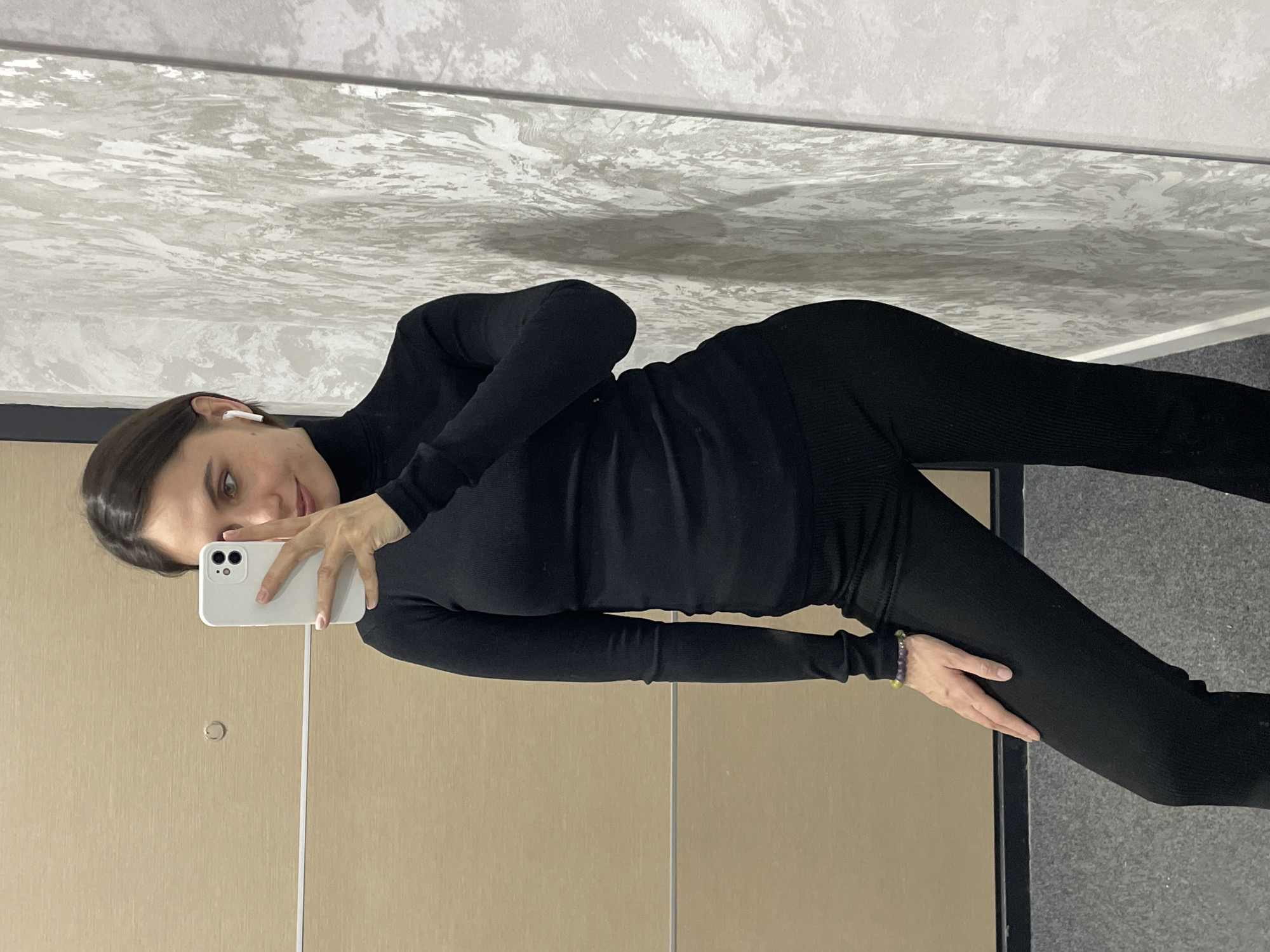 A female subject stands in a mirror reflection, capturing a full-body selfie in an indoor setting in Izhevsk, Russia. She has dark hair, light eyes, and wears a black ribbed turtleneck top and matching black ribbed trousers. A white wireless earbud is visible in her right ear, and a multi-colored beaded bracelet adorns her left wrist. Her right hand, with light pink polished nails, holds a white smartphone with a dual-lens camera, partially obscuring her lower face. Her left hand rests on her left thigh. The environment visible in the reflection includes a grey carpeted floor. To the left, a light brown paneled surface features a horizontal silver strip and a circular silver fixture. To the right, a textured grey wall displays abstract lighter grey and white patterns.