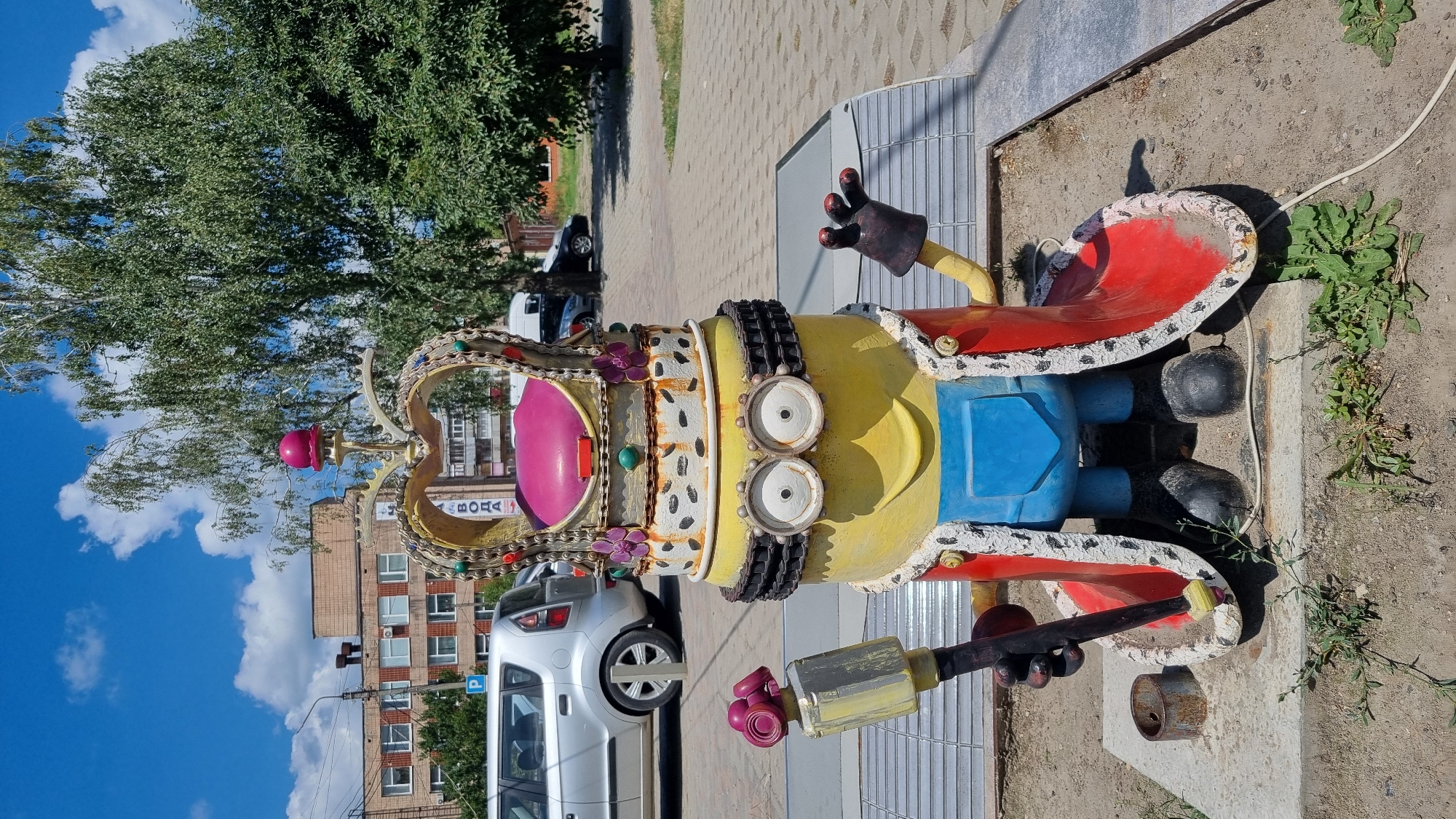 The image shows a statue of a Minion character dressed as a king, with a crown and scepter. The statue is located in the city of Smila, Ukraine, on a sidewalk next to a street. In the background, there are buildings, trees, and parked cars. The Minion statue is yellow with blue overalls, a red cape lined with white fur, and a crown with a pink jewel on top. It holds a scepter in its left hand and its right hand is raised. The statue appears to be made of concrete or a similar material and is weathered, with some dirt and wear visible on its surface. A silver car is parked to the left of the statue, and a building with windows is visible behind it. The sky is blue with some white clouds.