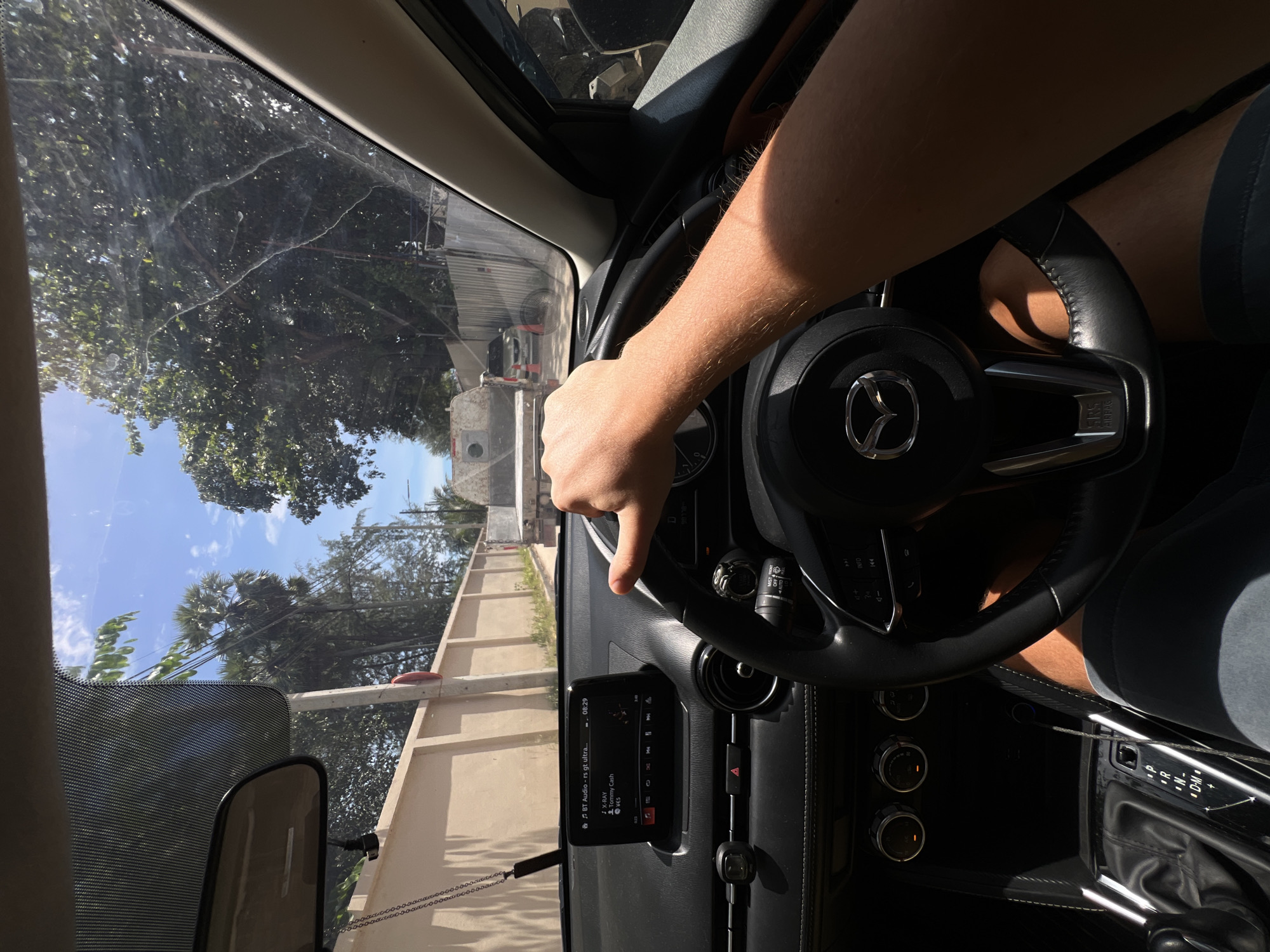 The image shows a first-person perspective from inside a car, with the driver's right hand on the steering wheel. The steering wheel is black leather with a silver Mazda logo in the center. The car's dashboard is visible, featuring a central infotainment screen displaying music information. To the left, the gear shift and climate control knobs are visible. Outside the car, a street is seen with a low beige wall on the left and lush green trees and palm trees on the right. Sunlight filters through the trees, casting shadows. In the distance, a white vehicle with a flatbed trailer and a small orange traffic cone are visible, along with other parked cars and a fence. The sky is blue with scattered clouds. The scene appears to be a sunny day in a tropical or subtropical urban environment.