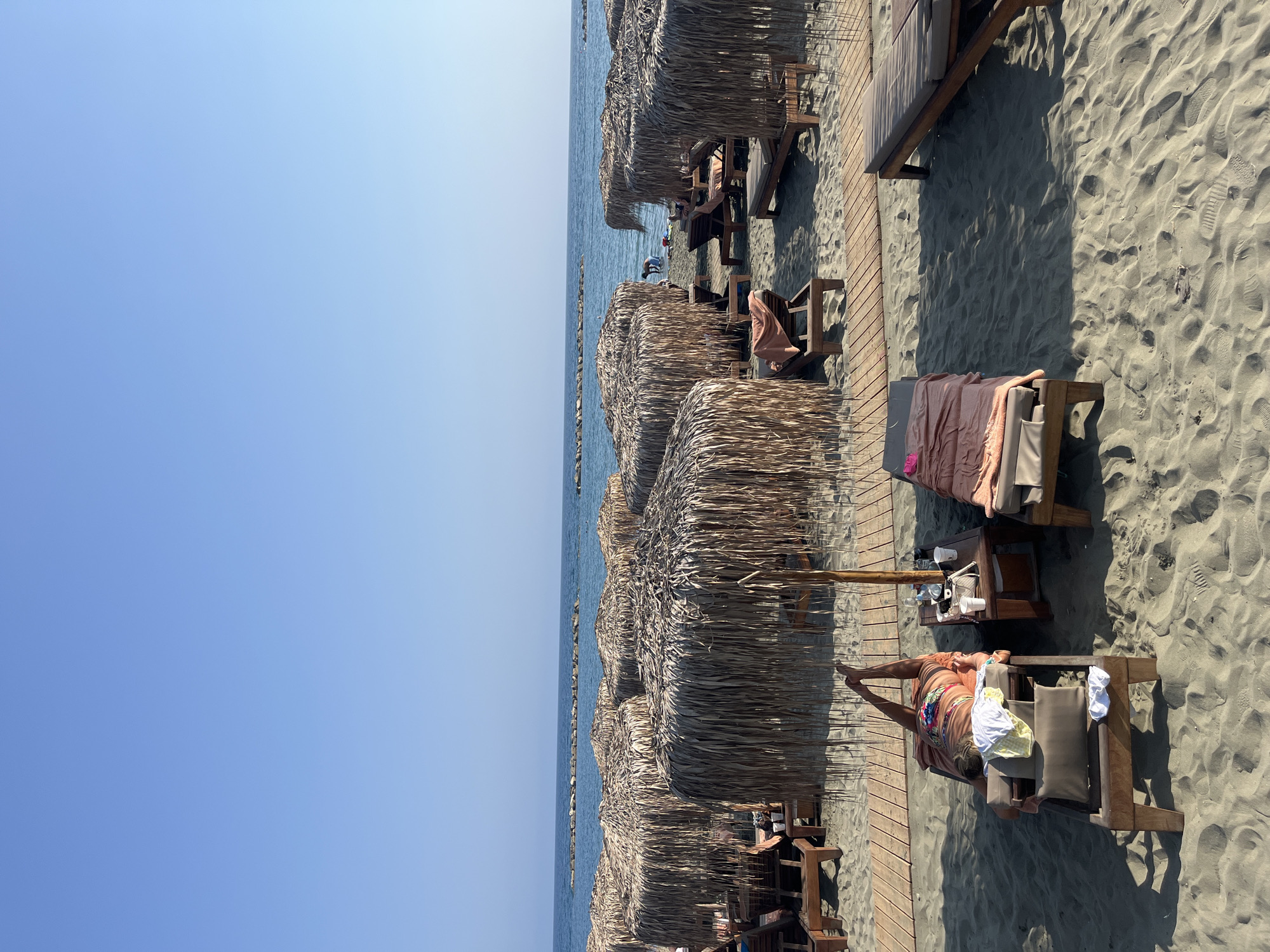 A wide-angle, eye-level shot captures a serene beach scene in Agios Tychona Community, Cyprus. The sand is a pale golden hue, with visible footprints and gentle waves lapping at the shore. In the foreground, several wooden lounge chairs are arranged on wooden pathways, some occupied by sunbathers. One person is lying on a lounge chair, wearing a colorful swimsuit and appearing to relax. Nearby, a small wooden table holds drinks and a bag. Behind these, a row of thatched umbrellas provides shade, their dry fronds creating a natural, rustic aesthetic. Beyond the umbrellas, the blue ocean stretches to the horizon, punctuated by a line of rocks that create a natural breakwater. The sky is a clear, bright blue, indicating a sunny day. In the distance, a few figures are visible near the water, suggesting other beachgoers enjoying the coastal atmosphere. The overall impression is one of peaceful relaxation and summer leisure.