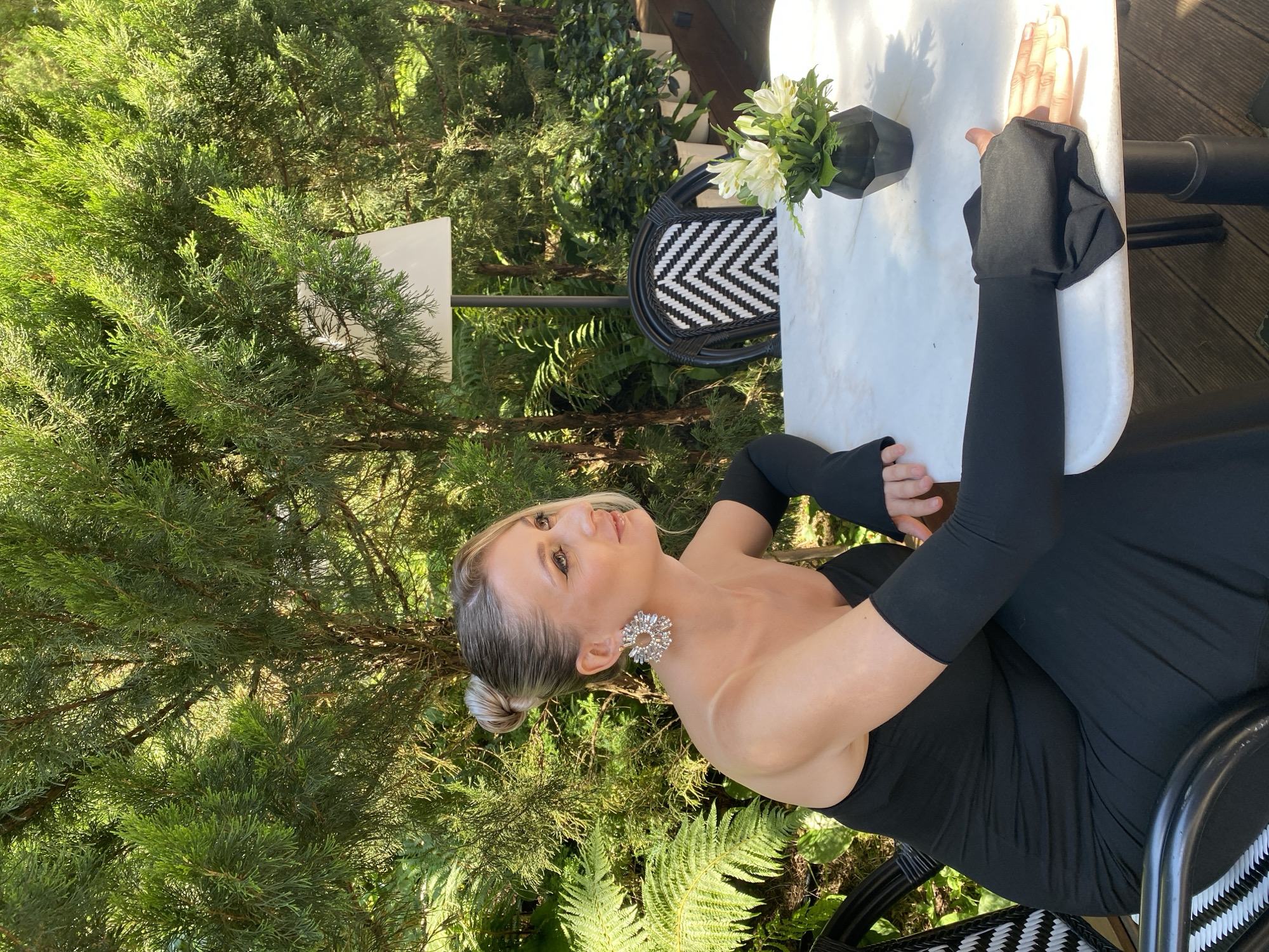 In Sochi, Russia, a blonde woman with her hair in a bun sits at a white table outdoors. She is wearing a black strapless dress and black fingerless gloves. She has large, sparkling earrings and her makeup is visible. The table is set with a black vase containing white flowers and greenery. Behind her, a chair with a black and white chevron pattern is visible. The background is filled with lush green trees and foliage, suggesting a garden or outdoor seating area. The lighting indicates it is daytime.
