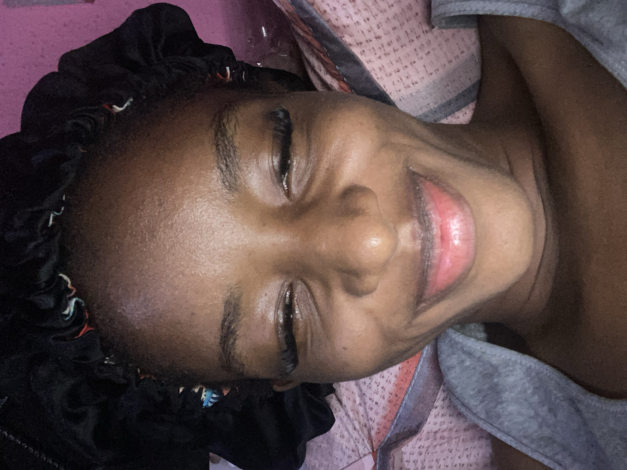 A close-up, head-and-shoulders selfie of a young Black woman with her eyes closed and a wide smile. She is wearing a black satin bonnet with colorful stitching. She is also wearing a grey tank top. The background appears to be a bed with pink and white patterned sheets. The lighting is soft and warm. There is no information about the location in Jos, Nigeria.