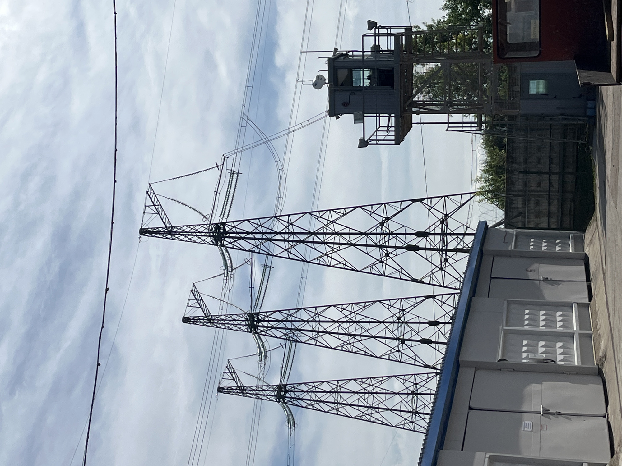 The image captures an industrial scene featuring large power transmission infrastructure under a partly cloudy daytime sky. In the foreground and midground, three prominent metal lattice transmission towers (electricity pylons) stand tall, supporting numerous power lines and insulators. These lines stretch across the sky, connecting to the towers.
To the right, an elevated, grey metal control cabin or guard post is visible, featuring windows and equipped with what appear to be external lights and a spherical white sensor or antenna. Further to the right and slightly behind, part of a reddish-brown structure, possibly another cabin or part of a larger building, is seen. Below these structures, a row of modular, light-grey utility boxes or containers with blue roof trim lines the ground, likely housing electrical equipment. These boxes have multiple doors with visible handles and locks, and some show corrugated or textured panels.
The background includes dense green foliage and trees, suggesting the area borders a natural or vegetated space. The ground on the far right appears to be paved with asphalt or concrete. The lighting indicates it is daytime, with bright but diffuse sunlight, and the sky is a pale blue with scattered clouds. The overall scene suggests a functional area related to power generation or distribution. On the red-colored structure, faint Cyrillic text reading "ОХРАНА" (meaning "GUARD" or "SECURITY") is partially visible above a window.