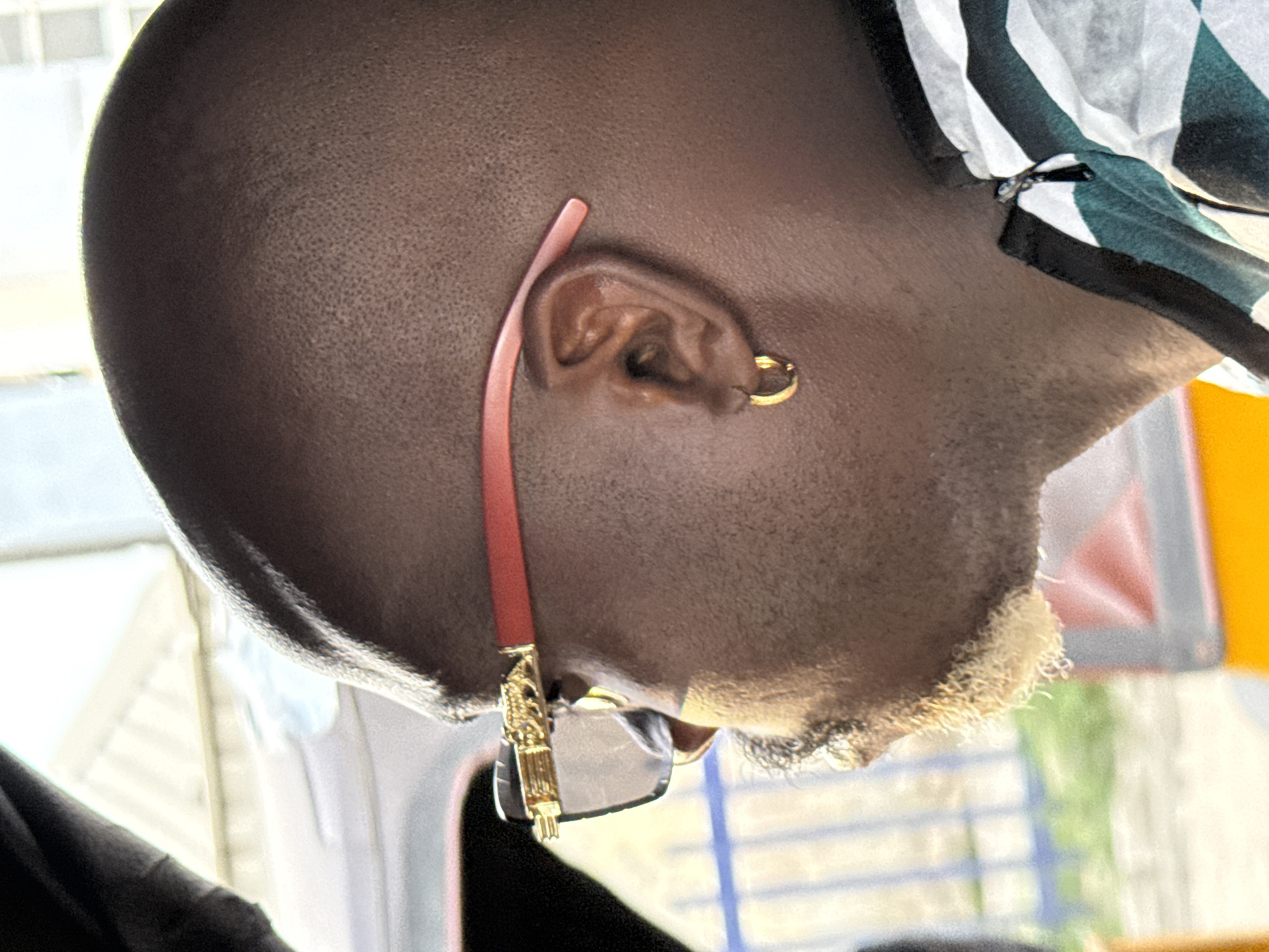 An extreme close-up, rotated 90 degrees clockwise, shows a man's head, ear with a gold hoop earring, a portion of sunglasses with red frames and gold decorative accents, and part of a striped head covering. The man has very short hair on top and dyed blond hair on his lower face. The specific location in Ikorodu, Nigeria, is not discernable from the available image.
