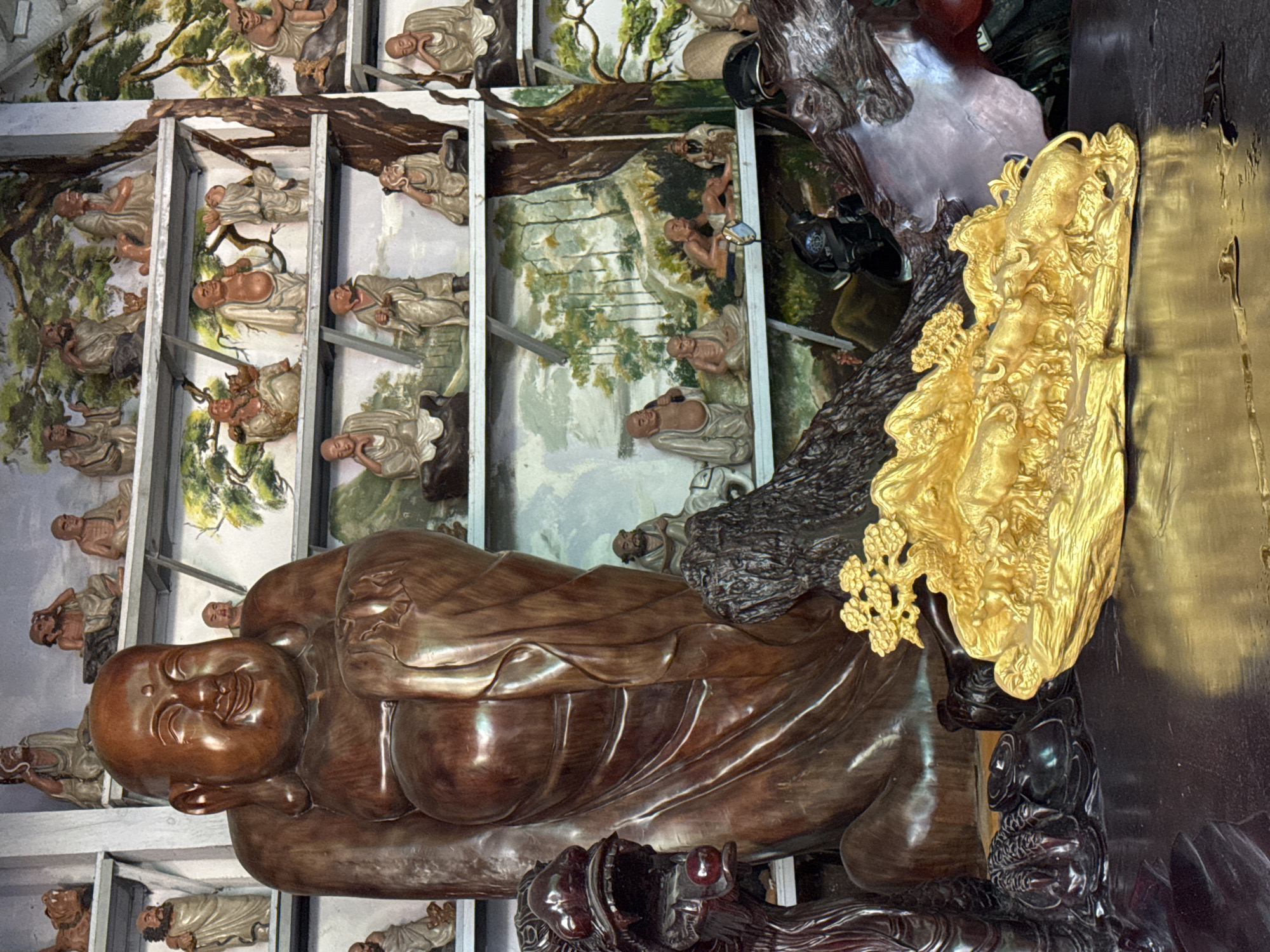 The image shows a collection of religious and decorative statues arranged on shelves in what appears to be a temple or shrine. Dominating the foreground is a large, smooth, reddish-brown wooden statue of a seated, smiling Buddha. To the left of the Buddha, a dark wooden dragon head statue with its mouth open and a red orb in its mouth is visible. To the right of the Buddha, a dark, textured wooden sculpture is present. In front of the large Buddha statue and partially obscured by it is a golden sculpture of several bulls on a rocky landscape with a small tree. The background consists of multiple shelves adorned with smaller statues of Buddhas and robed figures, some of which are placed in front of painted murals depicting landscapes and trees. The overall scene suggests a devotional or cultural space. This specific scene is located in a temple in Xuan Thanh village, Ninh Binh province, Vietnam.