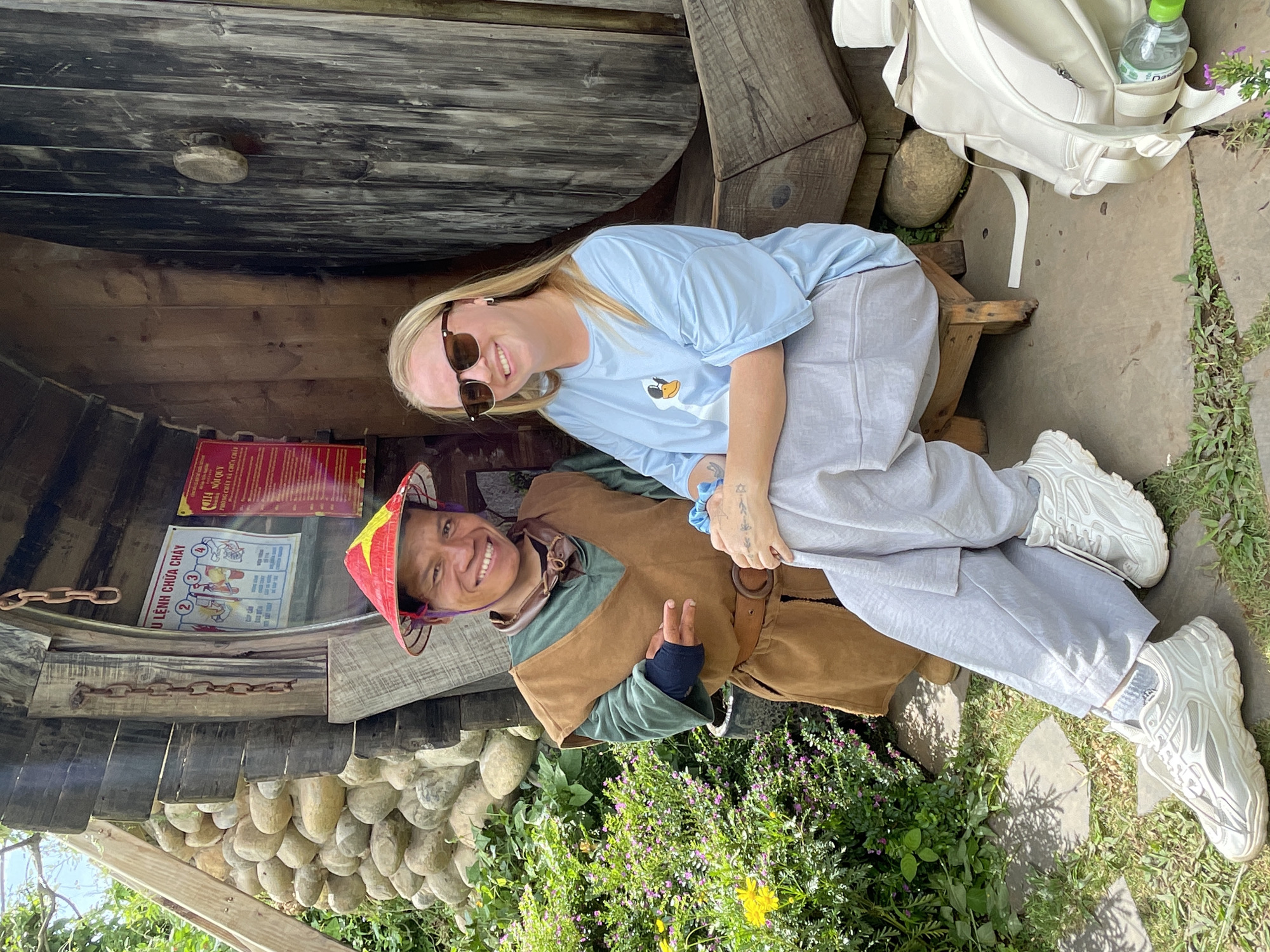 In Thôn 1, Vietnam, two individuals are photographed outdoors in front of a wooden structure. A man, wearing a conical hat adorned with red and yellow stars, a brown vest over a green garment, and a leather belt, is positioned on the left. He has a wide smile and appears to be gesturing with his left hand. To his right, a woman with blonde hair, sunglasses, and a light blue t-shirt with a duck graphic is seated on a wooden stool. She is smiling and has tattoos on her left forearm. A white backpack and a water bottle with a green cap are visible on the ground near her. The background features a rustic building constructed with stones and wooden planks, and lush greenery with purple and yellow flowers.