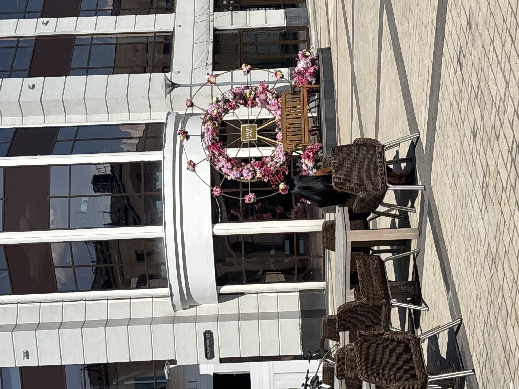This is an exterior, eye-level, medium shot of a plaza with outdoor seating and a decorative floral display in front of a building. The building is modern with large, dark-tinted windows and light-colored facade. To the left of the building entrance is a bicycle. In the foreground, several tables with woven chairs are arranged on a paved area. A woman with long dark hair is seated at one of the tables, facing away from the camera. Behind the tables, a large circular floral arrangement with pink and white flowers is mounted on a metal frame. A wooden bench is placed in front of the floral display, and a sign reading "ESTATE MALL" is visible within the circle. The ground is paved with square tiles, and shadows from the tables and chairs are cast across the plaza. A street lamp is visible to the right of the floral display. The overall impression is of a pleasant, sunny day in an urban public space.