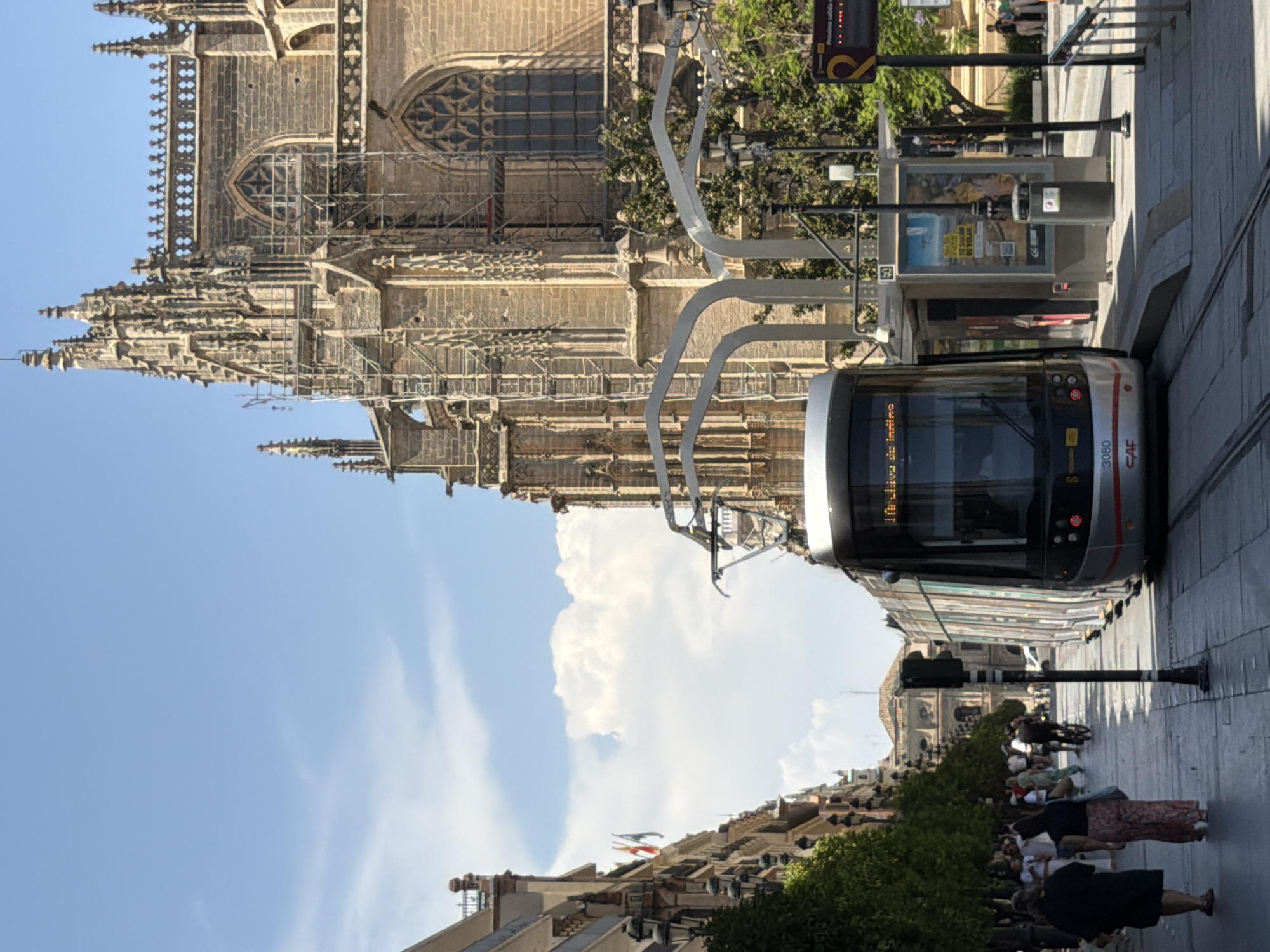 In Seville, Spain, a modern tram travels down a street lined with trees and historic architecture. The tram, with its silver and white exterior and "CAF" logo visible, is positioned on tracks in the foreground. The street itself is paved with large stone tiles and features a sidewalk with people walking and standing. To the right of the tram is a modern tram stop shelter with advertisements displayed. Looming in the background on the right is a large, intricate Gothic-style building, likely a cathedral or religious structure, partially covered in scaffolding, suggesting ongoing renovation. On the left side of the street, a row of buildings with balconies and lampposts adds to the urban landscape. The sky above is a clear blue with scattered white clouds. The scene captures a moment of daily life in Seville, blending its historic charm with its modern transportation infrastructure.
