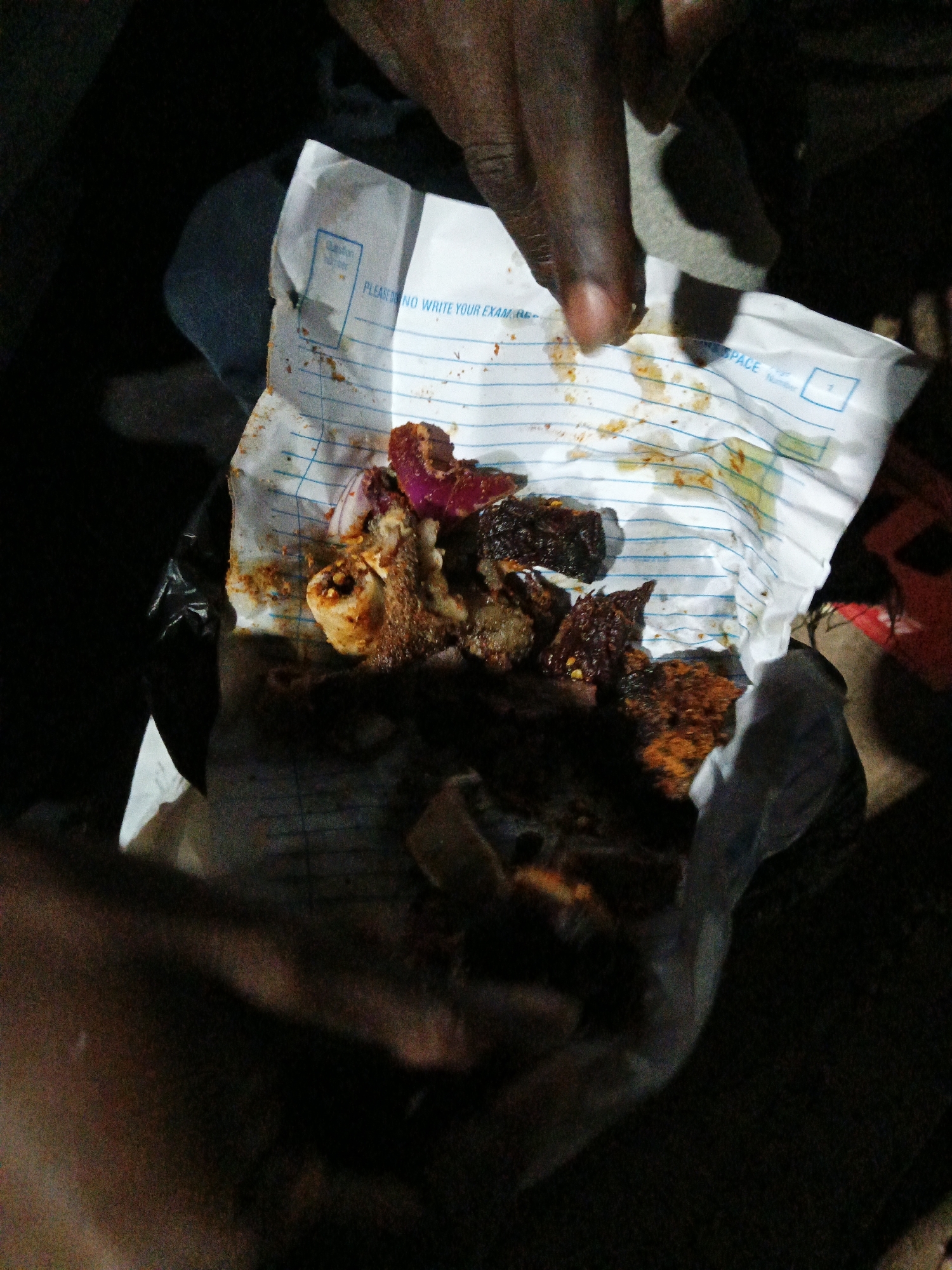 In Bukuru, Nigeria, a close-up shot captures a person's hands holding crumpled paper containing food. The paper, bearing printed lines and text, is soiled with food remnants. The food itself appears to be a mix of meat, onions, and spices. The lighting is dim, suggesting it may be evening or nighttime, and the background is dark and indistinct, with some hints of fabric or clothing. The overall scene evokes a sense of casual consumption of food, possibly from a takeaway or street vendor.