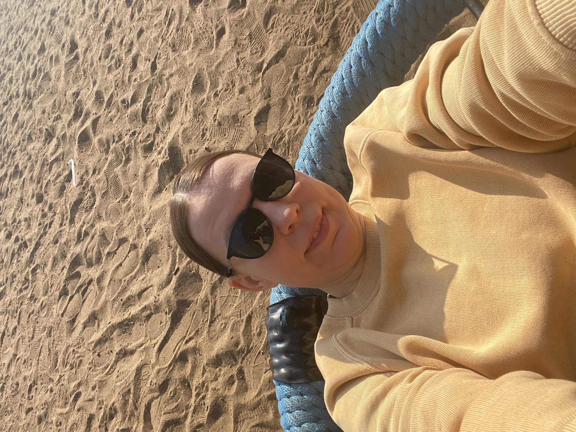 A person wearing sunglasses and a tan-colored sweatshirt lies in a blue swing located on a sandy surface, possibly a beach. A white straw sits on the sand. The location may be Chaikovsky, Russia.