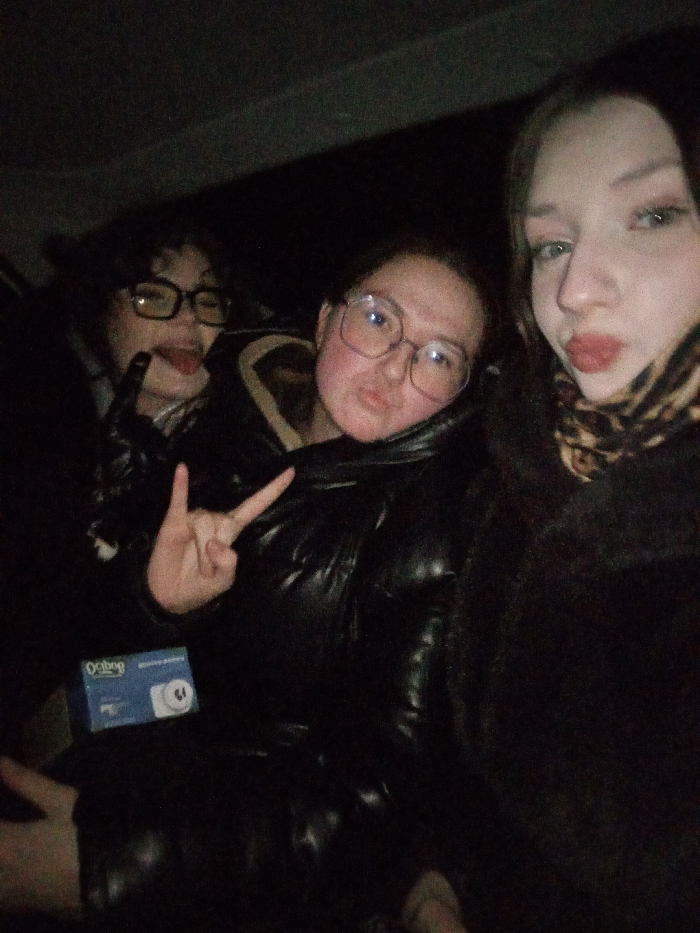 The image captures three young females in a dimly lit, enclosed space in Urtalga, Russia. The individual on the left, with dark hair and glasses, extends their tongue. The central individual, wearing a dark puffer jacket and glasses, forms a peace sign with their right hand and has puckered lips. The individual on the right, with long dark hair, a dark furry garment, and a leopard print scarf, also presents puckered lips. A light blue box labeled "OcBag" with the number "15" is situated in the lower left foreground, next to the central figure. The background is indistinctly dark, consistent with an interior setting with low light.