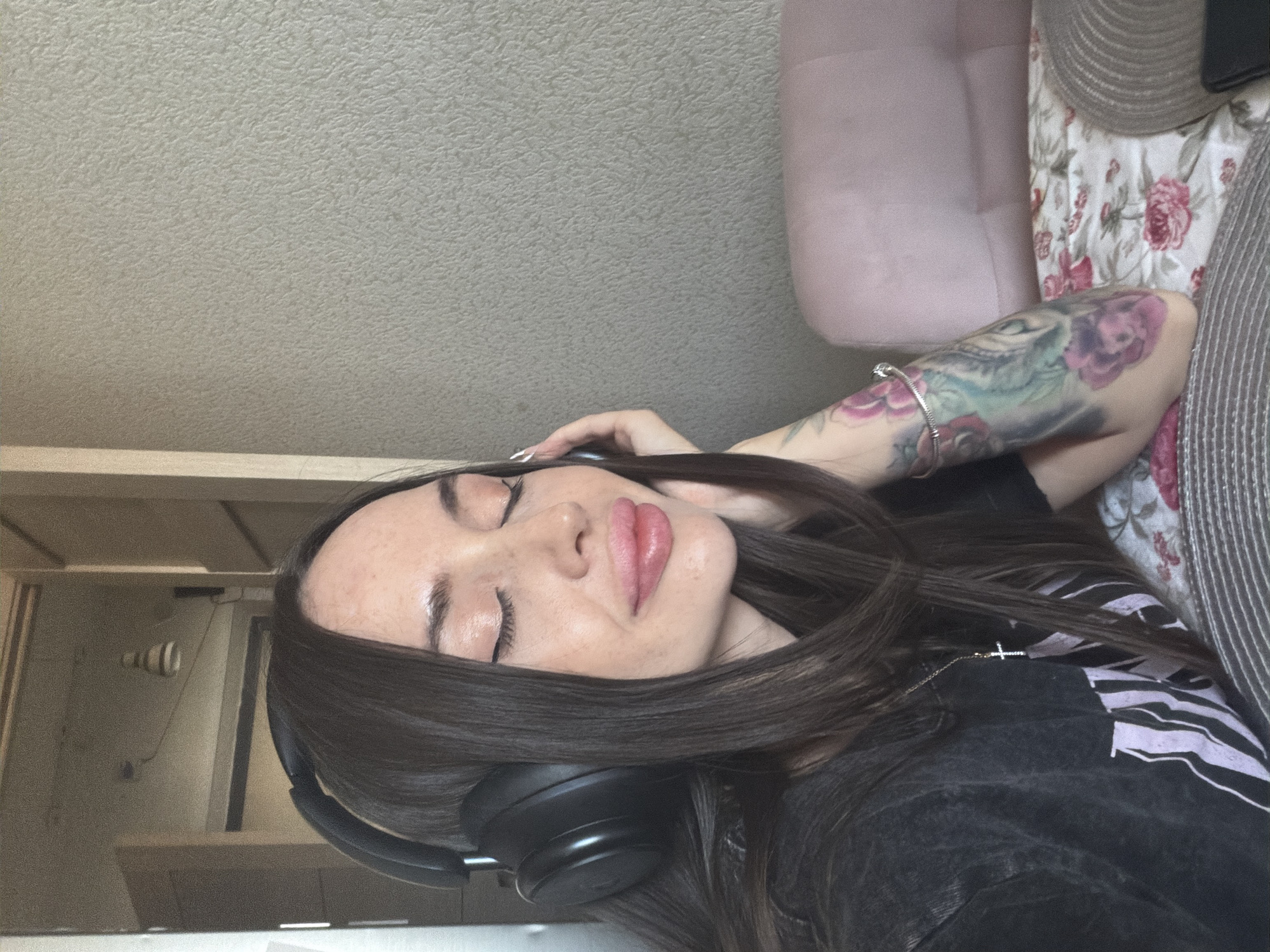 A woman is reclining with her eyes closed, wearing black headphones and a black shirt. Her dark hair is spread around her face. She has a tattoo on her left arm. The background includes a wall, a couch, and items on a surface, one of which appears to be a hat. The location is not discernible from the image.
