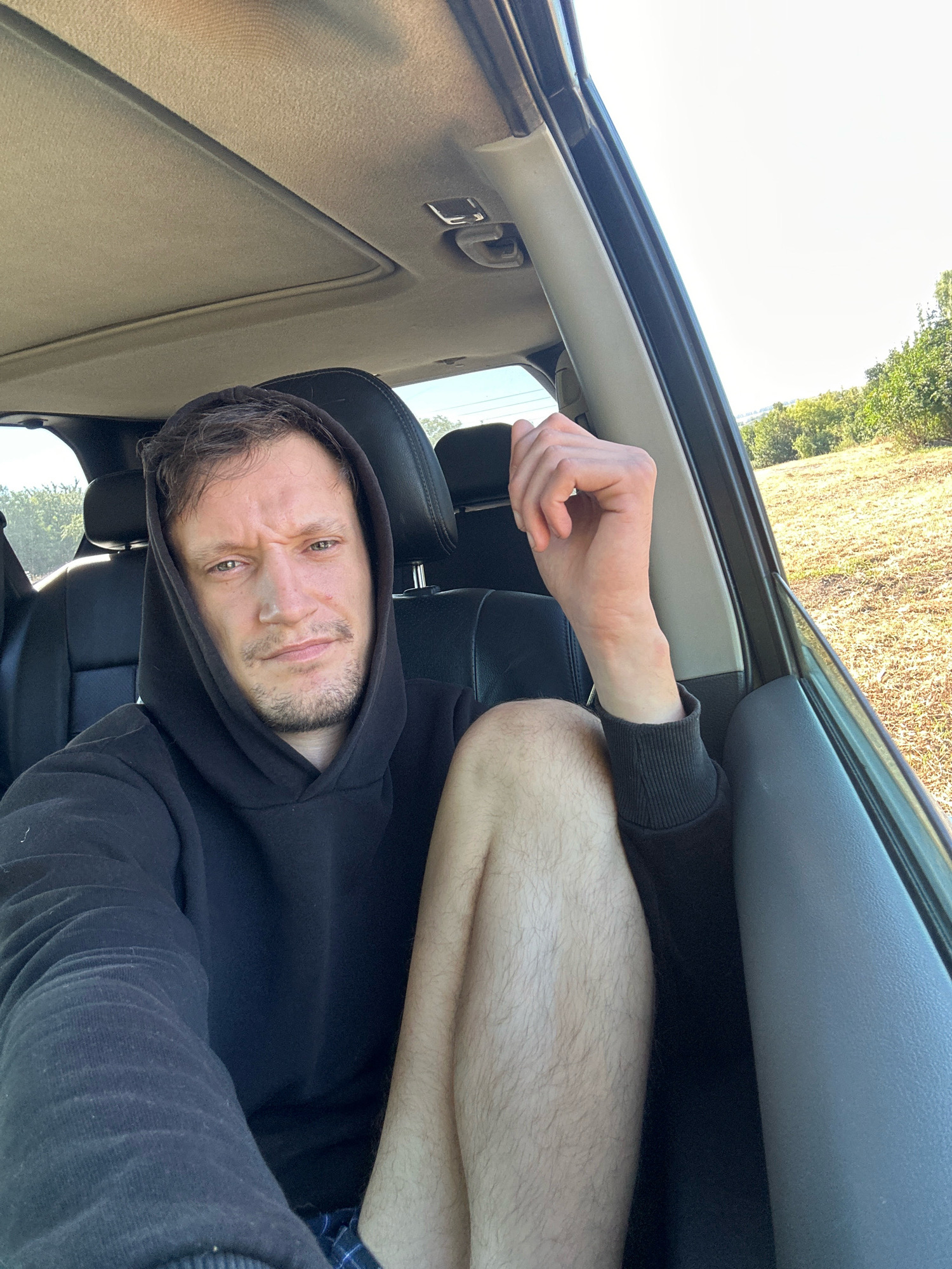 The image shows a man with light brown hair and a short beard, wearing a black hoodie, sitting inside a car. He is looking at the camera with a slight frown. His right arm is resting on his leg, which is also visible and appears to be unshaven. The interior of the car is dark, with black leather seats and a sunroof. Outside the car, there is a dry, grassy field with trees in the background. The lighting suggests it is daytime. The location is identified as Makiv, Ukraine.