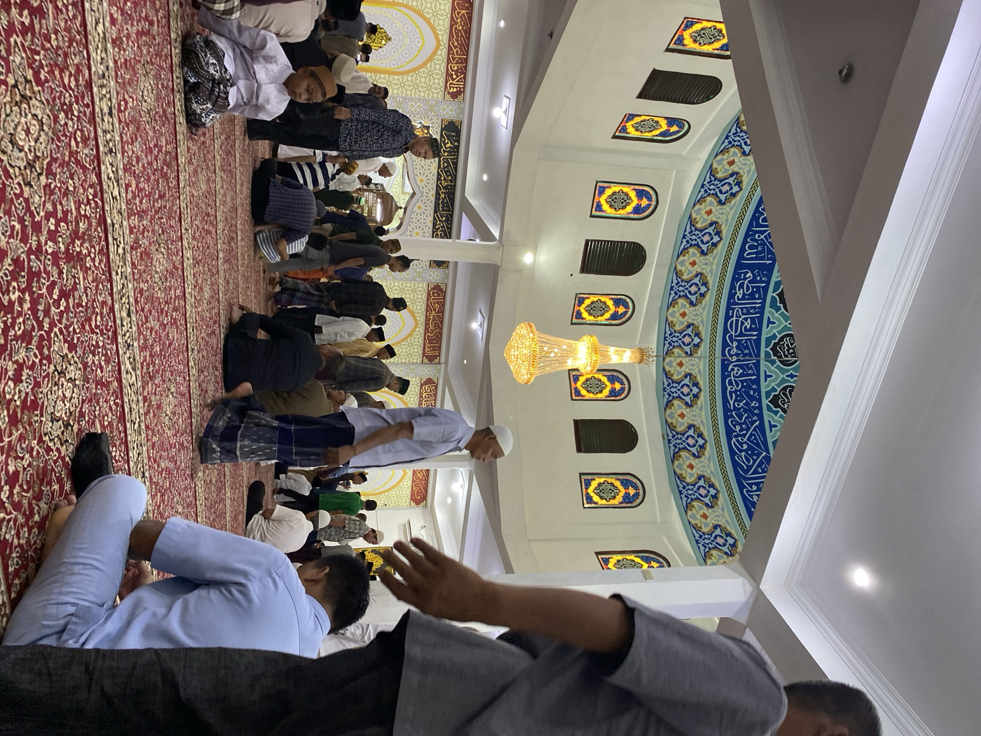 The image shows the interior of a mosque in Lhokseumawe, Indonesia, filled with a congregation of men and boys. The setting appears to be during or immediately before a prayer service, as many individuals are seated on a red carpeted floor, some in prayer positions. The mosque features a domed ceiling adorned with intricate Islamic calligraphy and colorful decorative motifs. Arched windows with stained glass patterns are visible along the walls. A large, ornate chandelier hangs from the center of the dome. The lighting in the mosque is a mix of artificial light from the chandelier and ceiling fixtures. People are dressed in traditional Islamic attire, including peci caps and sarongs. The overall atmosphere suggests a religious gathering.
