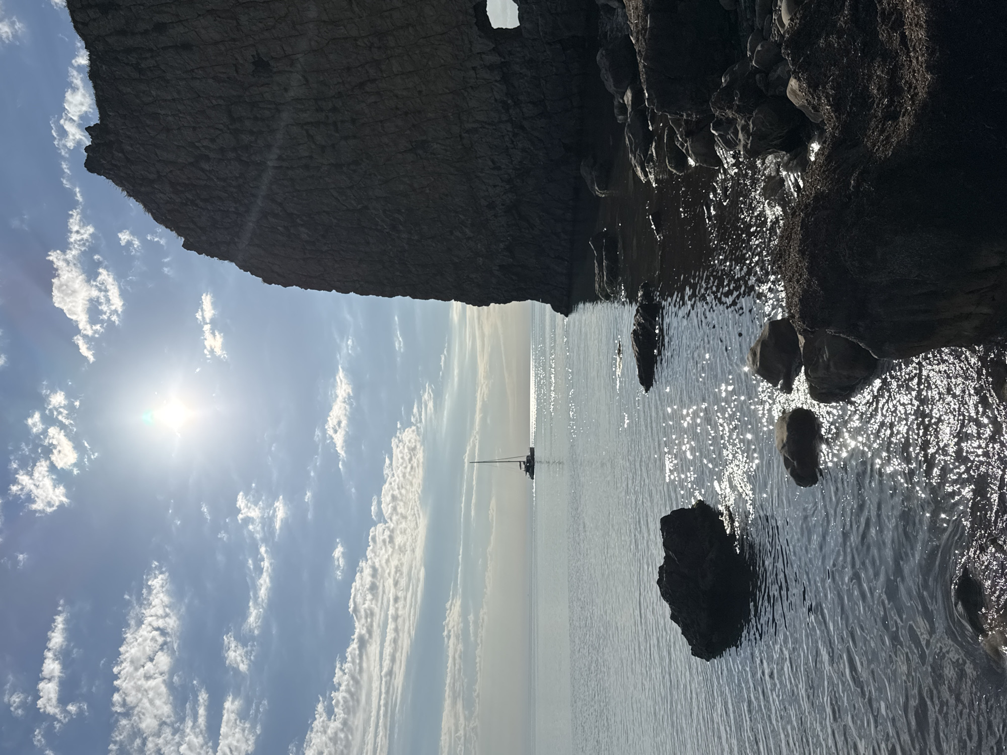 This media file presents a serene coastal landscape, captured on a bright, sunny day, likely during the morning or late afternoon. The scene is dominated by a massive, dark, textured cliff face that occupies the left side and top portion of the image. A small, natural arch or opening is visible in the upper part of the cliff.
To the right of the cliff, calm, dark water, presumably the sea or ocean, stretches towards the horizon. The water's surface is illuminated by direct sunlight, creating numerous sparkling reflections across its expanse. Several dark rocks are scattered along the water's edge and within the water on the right, some partially submerged.
In the distance, a small boat, appearing to be a sailboat or yacht with a mast, is silhouetted against the bright horizon, resting on the tranquil water. The sky above is a clear blue, adorned with scattered, wispy white clouds. The sun is a prominent feature in the upper left of the sky, emitting a strong light and lens flare, contributing to the sparkling effect on the water.
The overall impression is one of natural beauty and peace. There are no visible people or discernible text in the image. The location is Divnomorskoe, Russia.