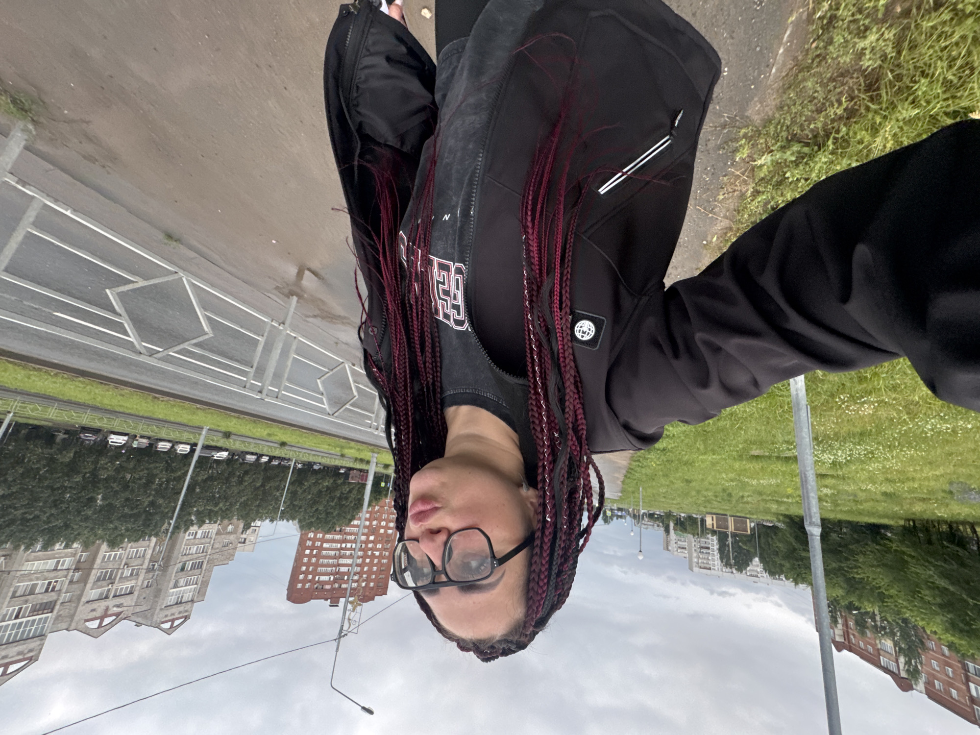 A woman with braided hair and glasses is taking a selfie outdoors in Tolyatti, Russia, on a cloudy day. She is wearing a black jacket and a grey t-shirt. The background shows apartment buildings, trees, a street with cars, and a sidewalk with a metal fence. The overall scene depicts an urban environment on a somewhat overcast day.