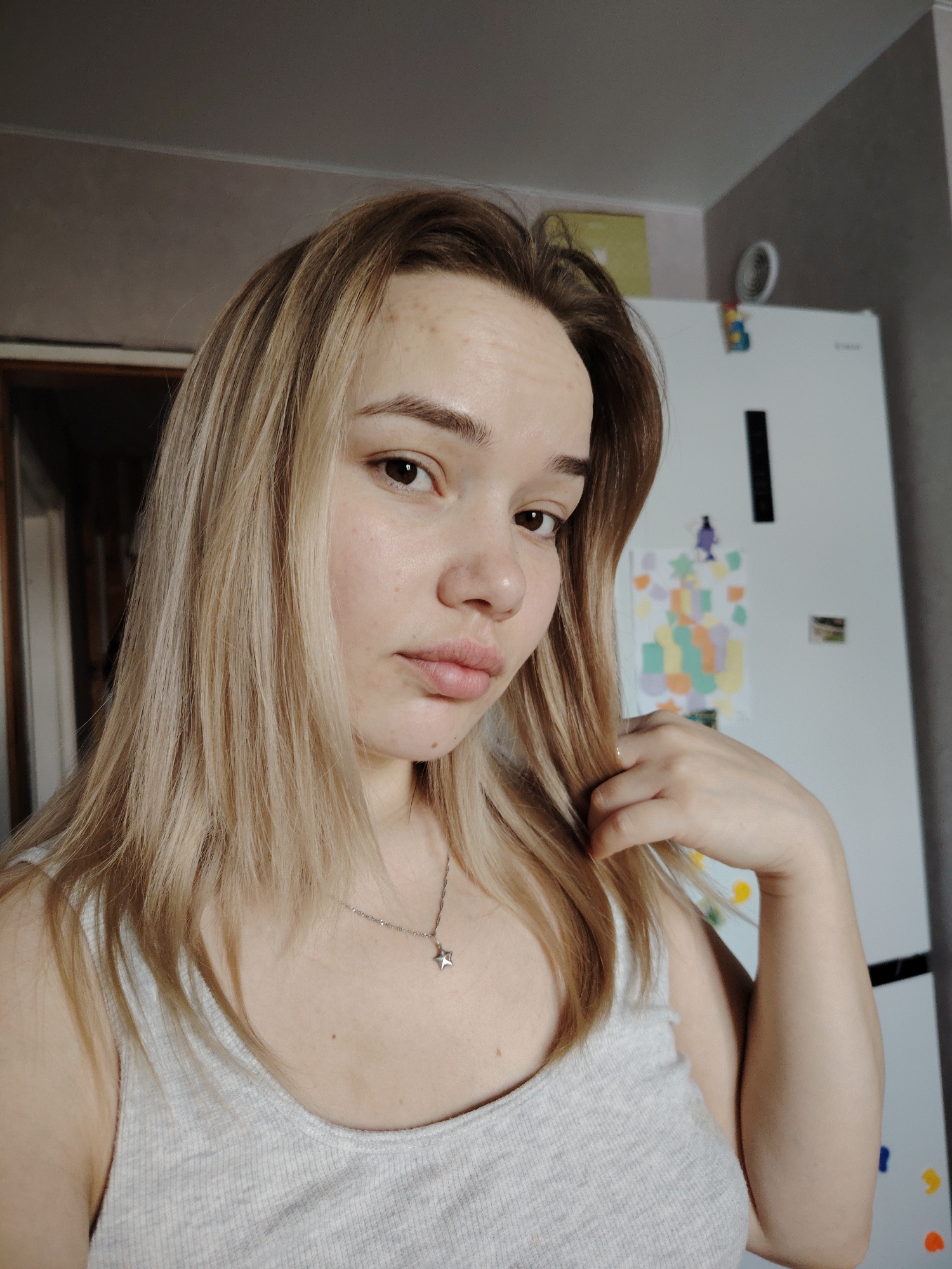 A young woman with blonde hair and fair skin is posing for a selfie indoors. She is wearing a light gray tank top and a silver necklace with a star pendant. Her hair is styled with dark roots and light blonde highlights. She has freckles on her forehead and cheeks. Her expression is slightly pensive. She is touching her hair with her right hand.
The background shows a typical domestic setting, possibly a kitchen. A white refrigerator is visible behind her, adorned with magnets, including what appears to be a child's drawing. There is also a doorway partially visible on the left side of the image.
There is no information in the image to confirm the location in the city of Марковский, Russia. The scene is generic and could be located anywhere.