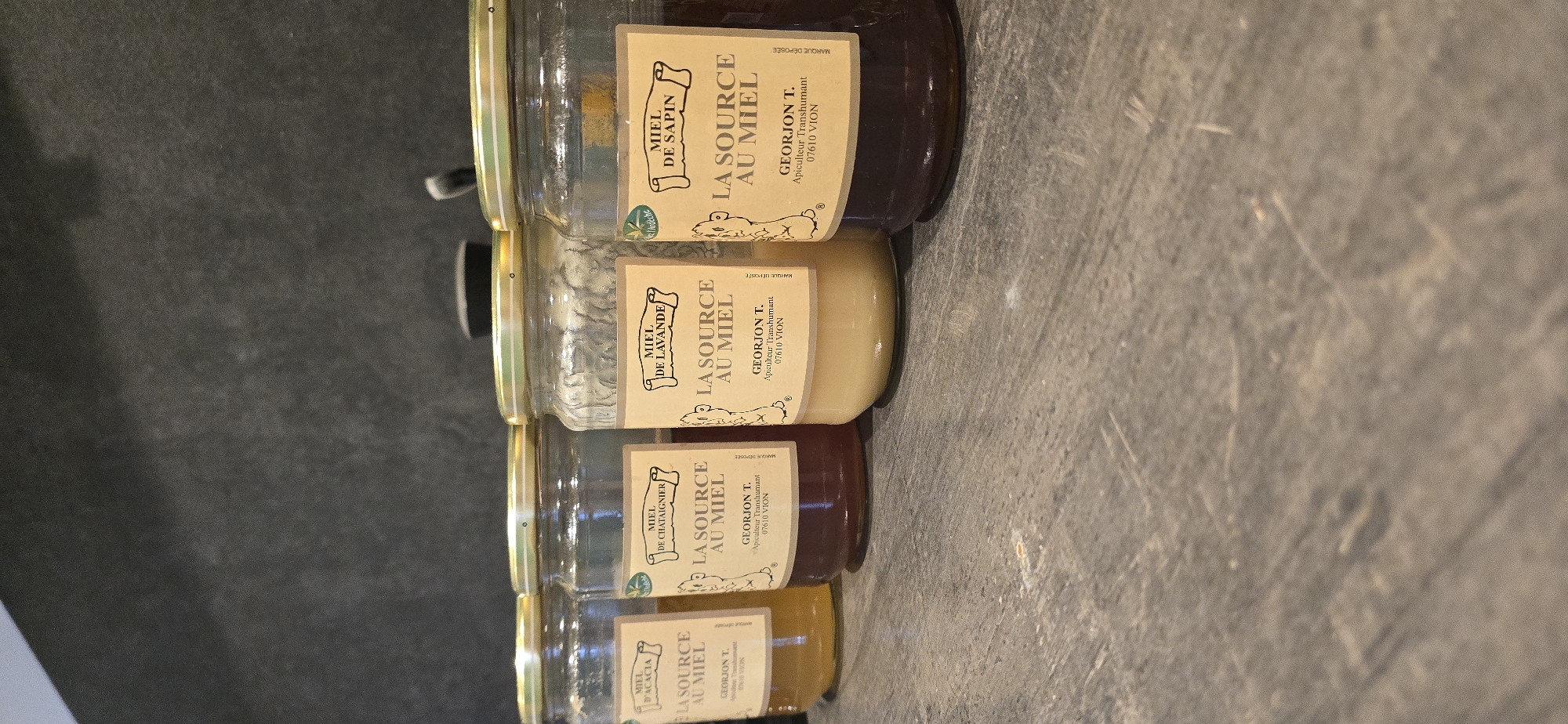 Four glass jars of honey are lined up on a dark, textured surface. Each jar has a golden lid and a light-colored label. The labels are all similar in design, featuring a drawing of a bee and text. From left to right, the labels read: "MIEL D'ACACIA LA SOURCE AU MIEL GEORJON T. Apiculteur Transhumant 07610 VION", "MIEL DE CHATAIGNIER LA SOURCE AU MIEL GEORJON T. Apiculteur Transhumant 07610 VION", "MIEL DE LAVANDE LA SOURCE AU MIEL GEORJON T. Apiculteur Transhumant 07610 VION", and "MIEL DE SAPIN LA SOURCE AU MIEL GEORJON T. Apiculteur Transhumant 07610 VION". The honey in the jars varies in color, from light yellow for acacia honey to dark brown for fir honey. The background is a dark, matte surface, possibly a wall. The image is taken from a slightly elevated angle, looking down at the jars. The overall impression is one of a product display, likely in a shop or market.