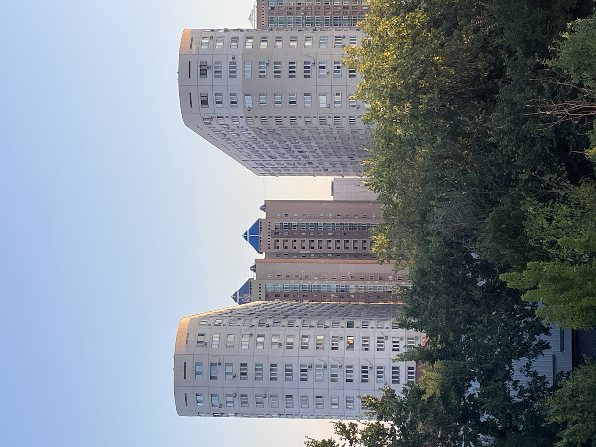 The image captures a view of apartment buildings in Almaty, Kazakhstan, under a clear blue sky. The foreground is dominated by dense green foliage from trees. In the mid-ground, three distinct apartment buildings rise above the trees. The building on the left is a tall, curved structure with a light-colored facade, featuring numerous windows and balconies. To its right is a narrower, taller building with a light brown and beige facade, capped with two distinctive blue pyramid-shaped roofs. Further to the right, another tall apartment building with a similar light-colored facade is visible, characterized by a repeating pattern of windows. The buildings appear to be residential complexes, indicative of urban development. The lighting suggests it is either early morning or late afternoon, with warm sunlight casting a glow on the tops of the buildings. The scene is tranquil and depicts a segment of Almaty's urban landscape.