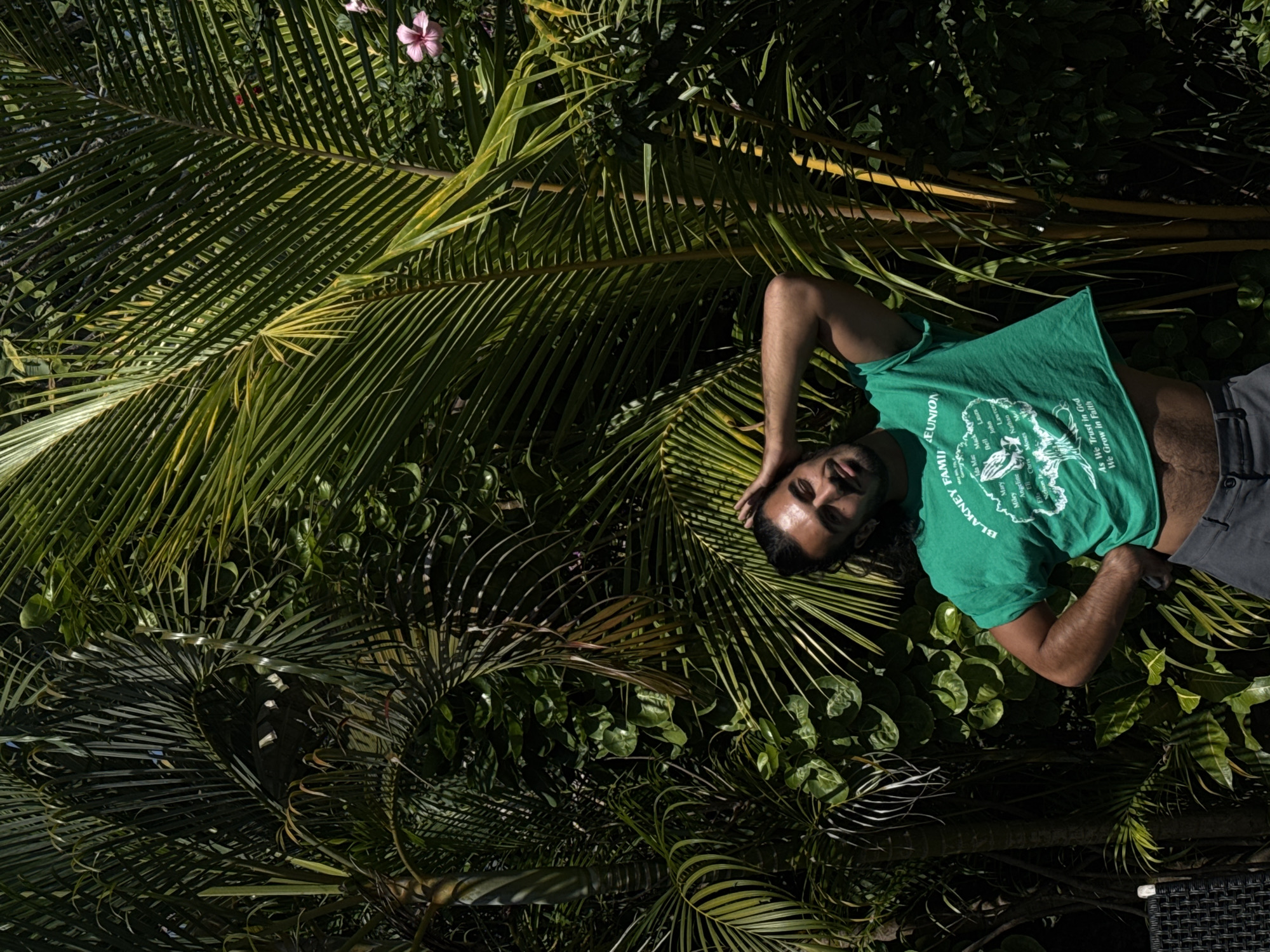 A man with dark hair and a beard is standing in front of lush green foliage and palm trees. He is wearing a bright green t-shirt that appears to be cropped, revealing his midriff, and gray pants. He has his right arm raised with his hand behind his head, and his left hand is on his hip. Sunlight filters through the leaves, creating dramatic shadows. A small pink flower is visible among the green leaves. The scene suggests a warm, tropical environment, potentially in North Kuta, Indonesia.