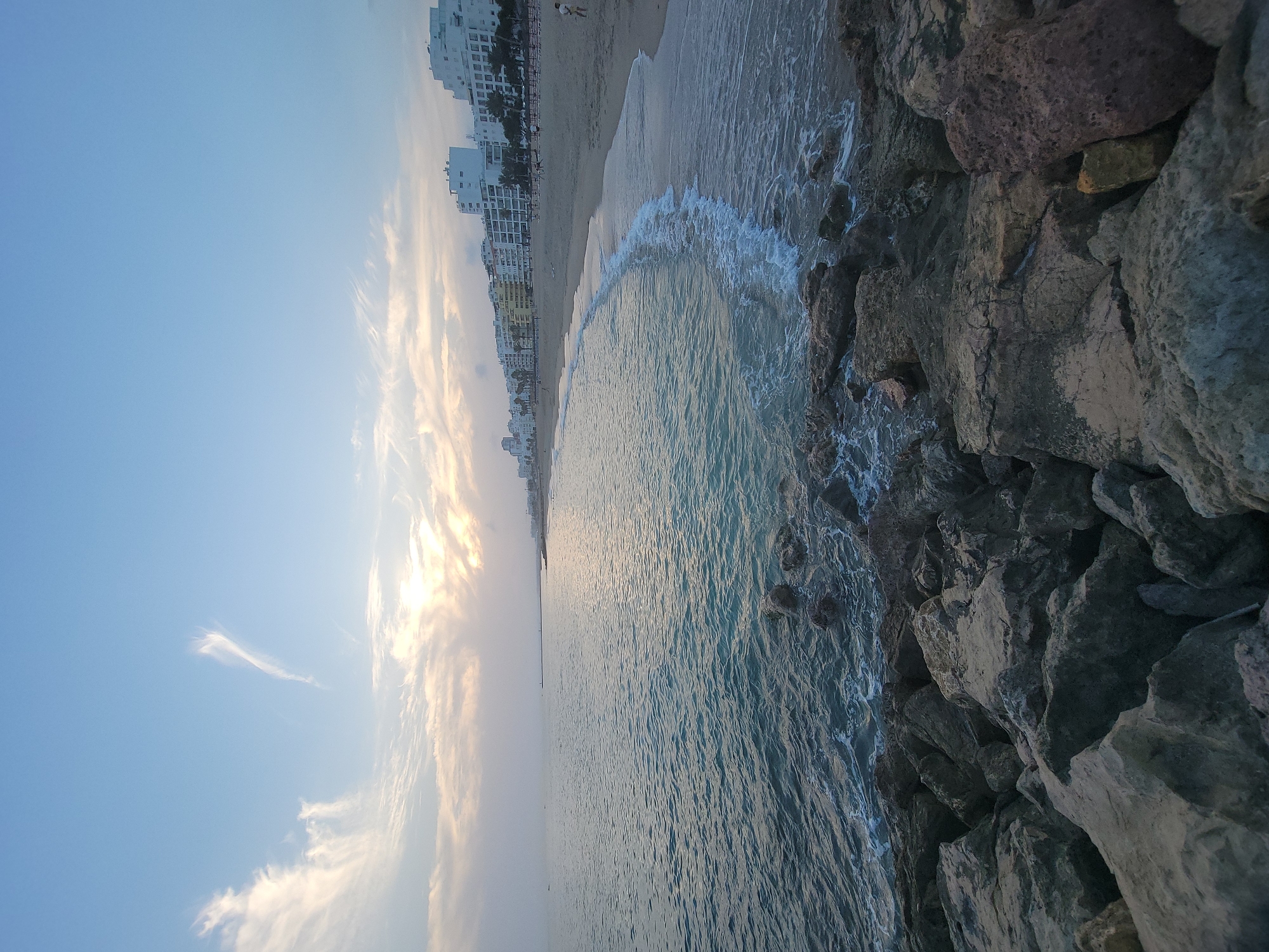 This is a wide-angle, low-angle shot of a rocky shoreline in Quarteira, Portugal, looking out at the ocean at dusk. The foreground is dominated by large, textured rocks of various shades of gray and brown. Beyond the rocks, the calm, turquoise ocean stretches to the horizon. Gentle waves lap at the shore, creating a white foam line.
To the right, a sandy beach is visible, lined with palm trees and a row of white and yellow buildings. The buildings are multi-story apartment or hotel complexes. A few scattered figures can be seen on the beach and near the water's edge.
The sky above is a pale blue, with soft, wispy clouds tinged with the warm colors of sunset. The sun itself is partially obscured by clouds, casting a soft, diffused light over the scene. The overall atmosphere is serene and peaceful.