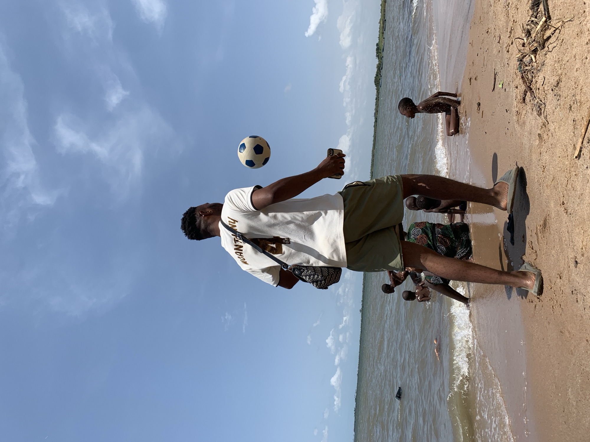 A man is standing on a beach juggling a soccer ball with his right hand while holding a mobile phone in his left. He wears a white t-shirt with the text "MCANTHONY" on the back, khaki shorts, and sandals. He is wearing a patterned shoulder bag. The beach appears to be in Ikorodu, Nigeria. Behind him and to his right, other people are playing and wading in the water. The sky is blue with scattered white clouds.