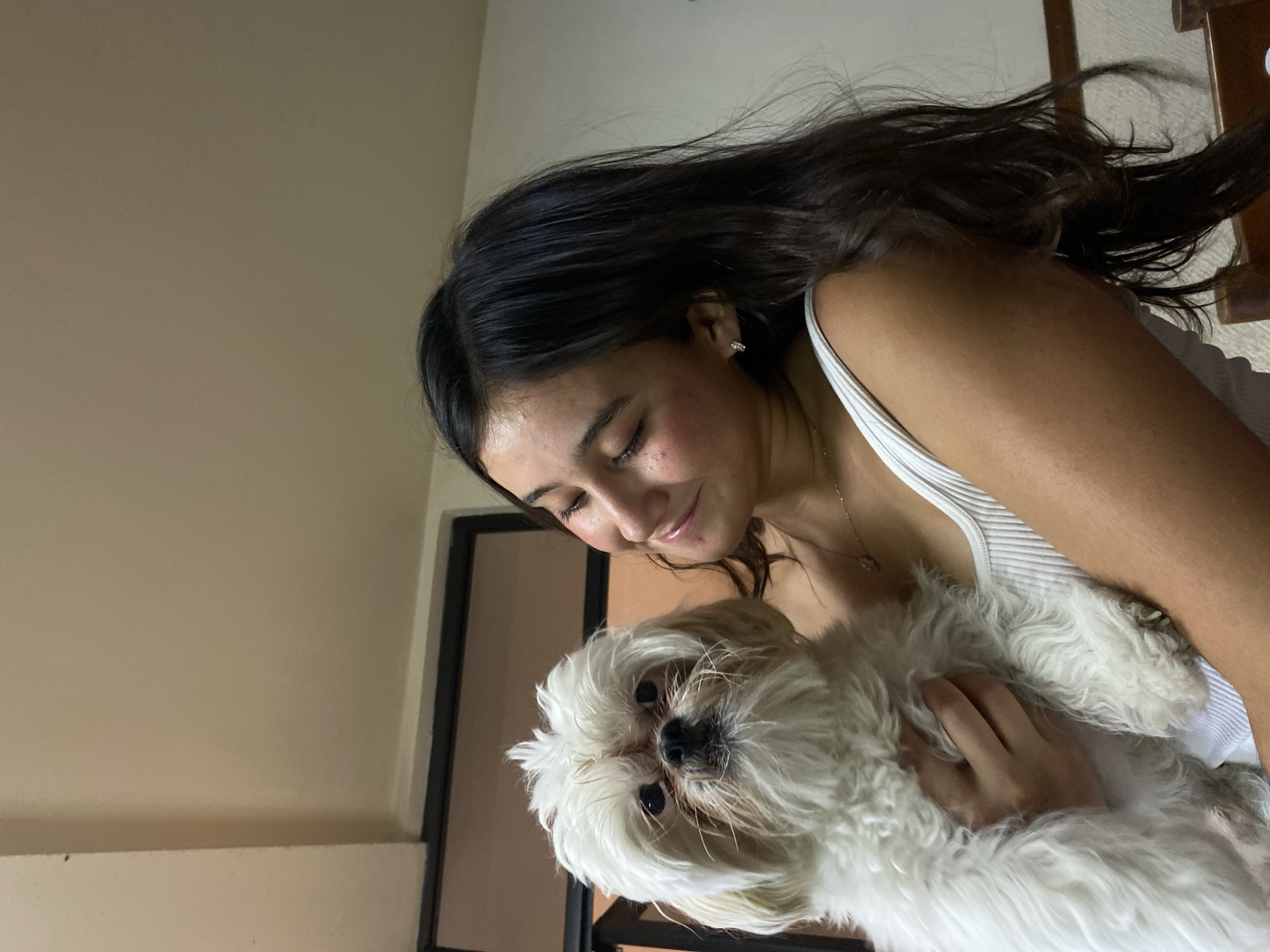 A medium shot captures a young woman indoors, holding a small, white, fluffy dog. The woman has long, dark hair and wears a white tank top. She looks down at the dog with a soft expression. The location appears to be a residence, indicated by the plain walls and a partial view of furniture in the background. The city and country are unspecified.