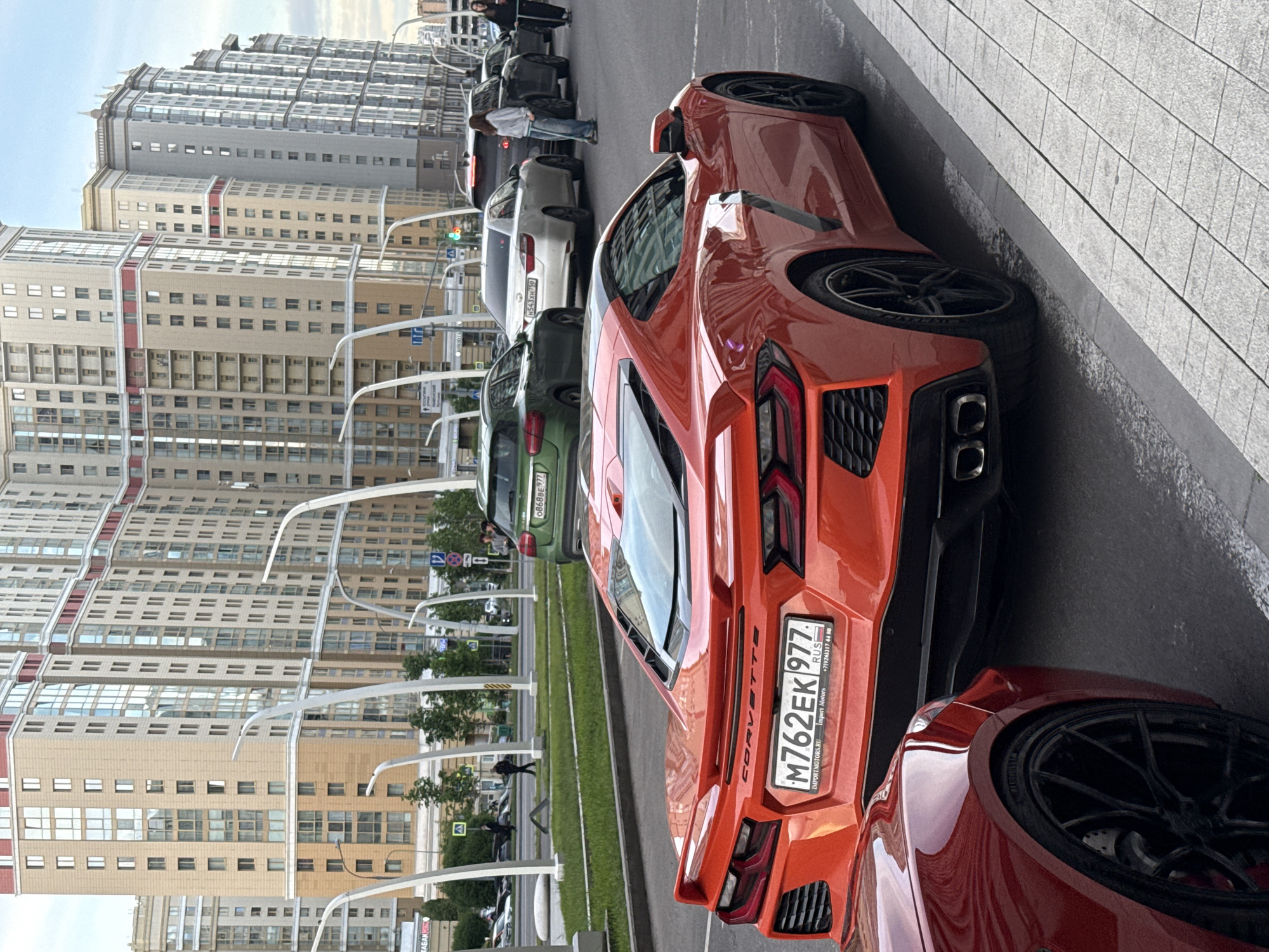 The image shows several cars parked along a street in what appears to be a city setting, likely Moscow, Russia, based on the license plates and Cyrillic text. The foreground features the rear end of an orange Corvette, with its distinctive taillights and the license plate "M762EK 977" visible. Next to it is another red sports car with a large black wheel visible. Behind the Corvette are other vehicles parked along the street, including a green sedan with the license plate "0868BE977" and a white sedan with "H543XH750" on the license plate. The background shows a row of tall residential buildings under a partly cloudy sky. The setting is a modern urban environment, with street lamps that have a modern design lining the street. People can be seen walking alongside the cars.