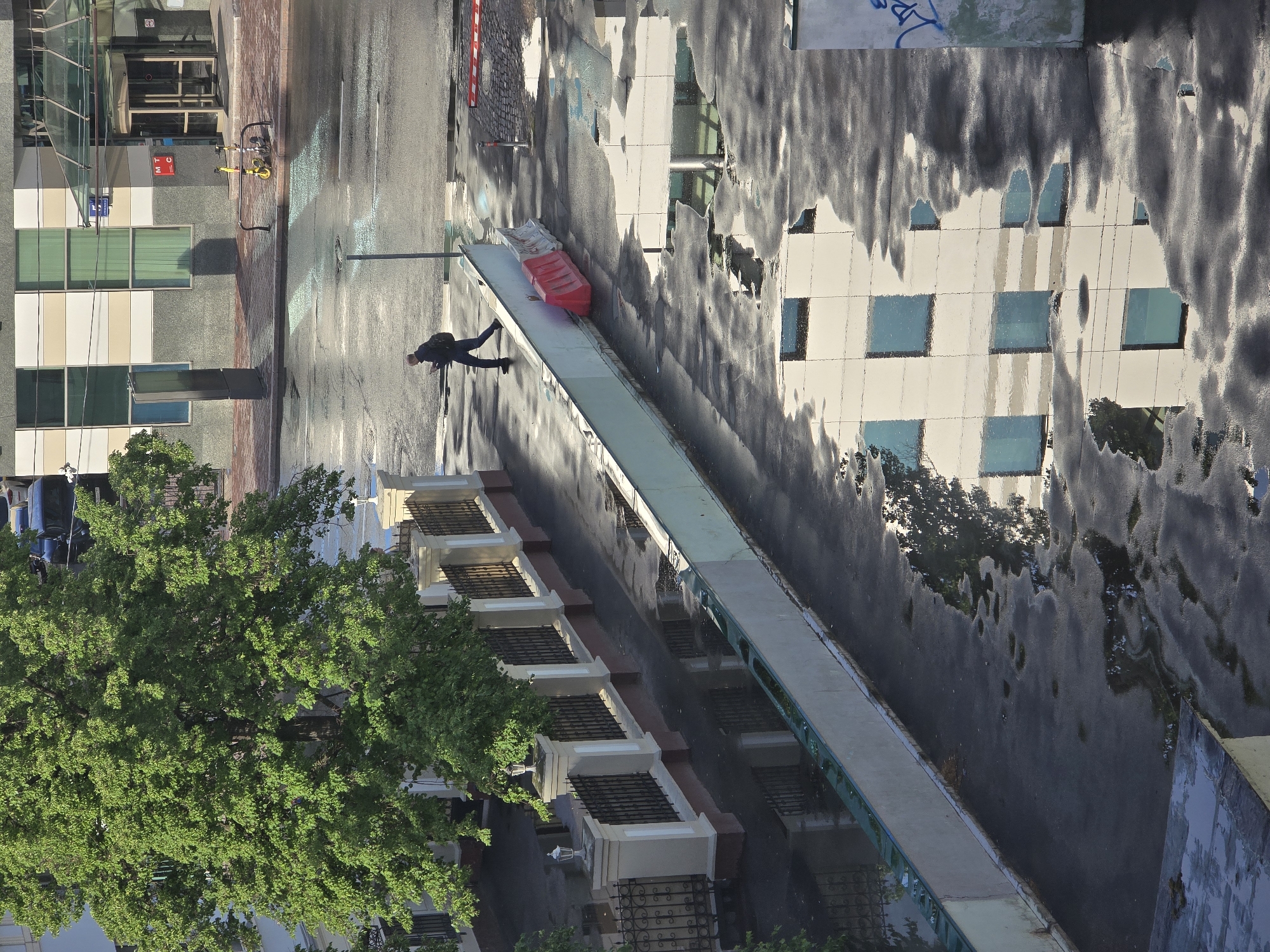 The image depicts an aerial view of a Moscow street scene in Russia. Rain is visible on the pavement, creating reflections of buildings and sky. A pedestrian in dark clothing is seen walking along the sidewalk. A pedestrian bridge with a red barrier is along the sidewalk. The area also features a mature tree with full green foliage, contributing to the urban landscape. Several buildings of various architectural styles are visible, including one with multiple windows reflecting the cloudy sky. The presence of street furniture and vehicles also indicates the urban environment.