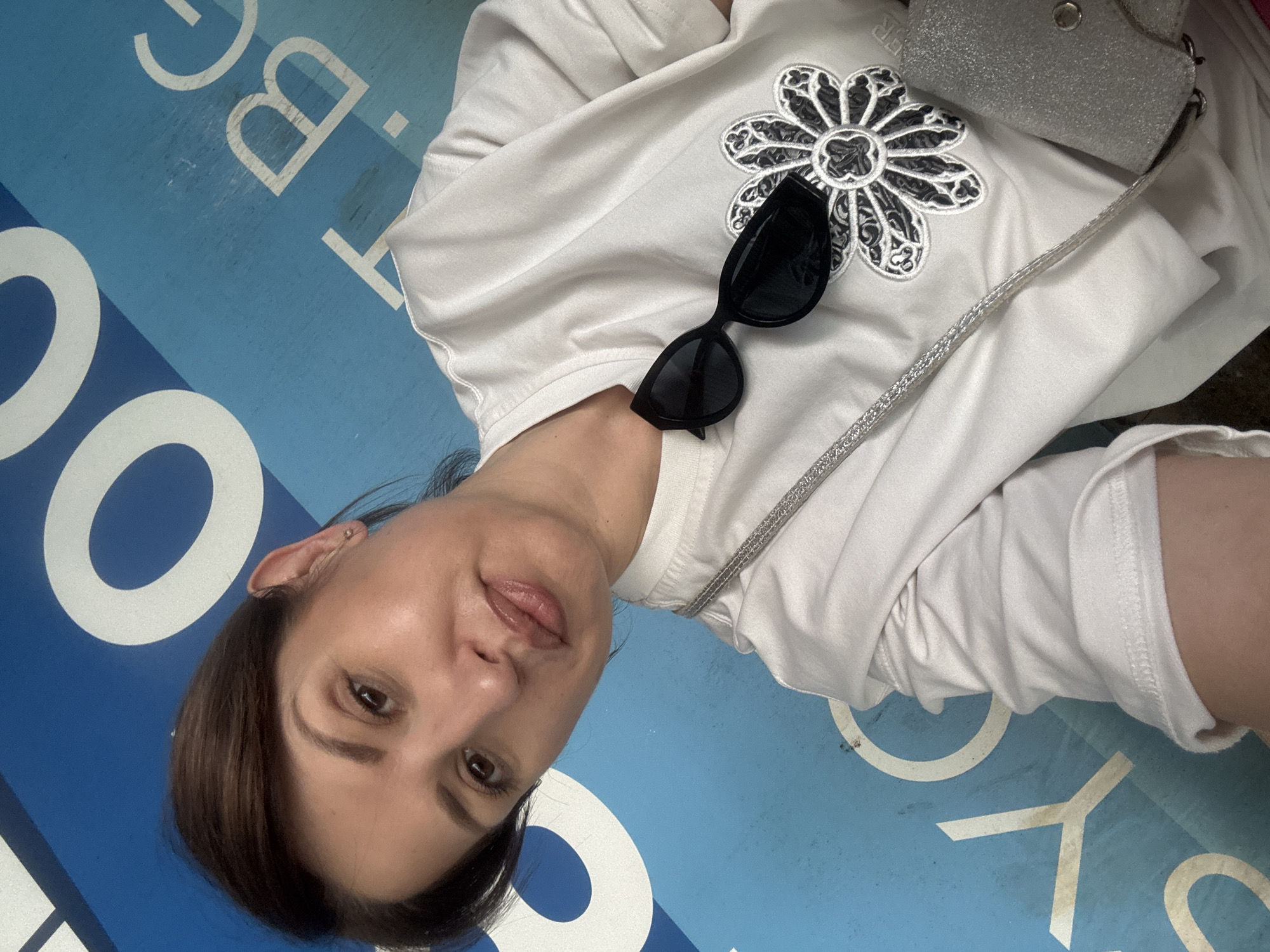 A woman with dark hair and light skin is pictured in a selfie. She is wearing a white shirt with a black and white flower design and has black sunglasses resting on the shirt. A silver purse with a chain strap is also visible. The woman is standing on a blue surface with white lettering, which reads "COOL BIKE SOFIA". This indicates the location is likely in Sofia, Bulgaria, where bike-sharing programs are present. The background is slightly blurred, suggesting a shallow depth of field.