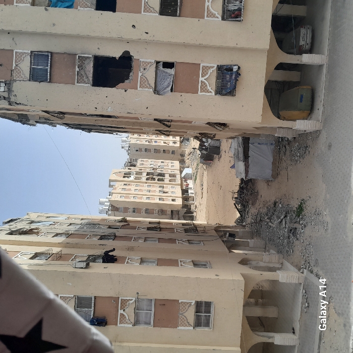 The image shows two buildings facing each other with a barren alleyway between them. The buildings are light brown with rectangular windows, some broken or covered with fabric. Debris litters the alleyway floor, with a makeshift shelter or tent visible in the distance. In the far distance, more buildings are seen under a clear sky. There is a glimpse of a road on the right. The image is tilted and appears to be taken from ground level, with a blurry object in the lower left corner.