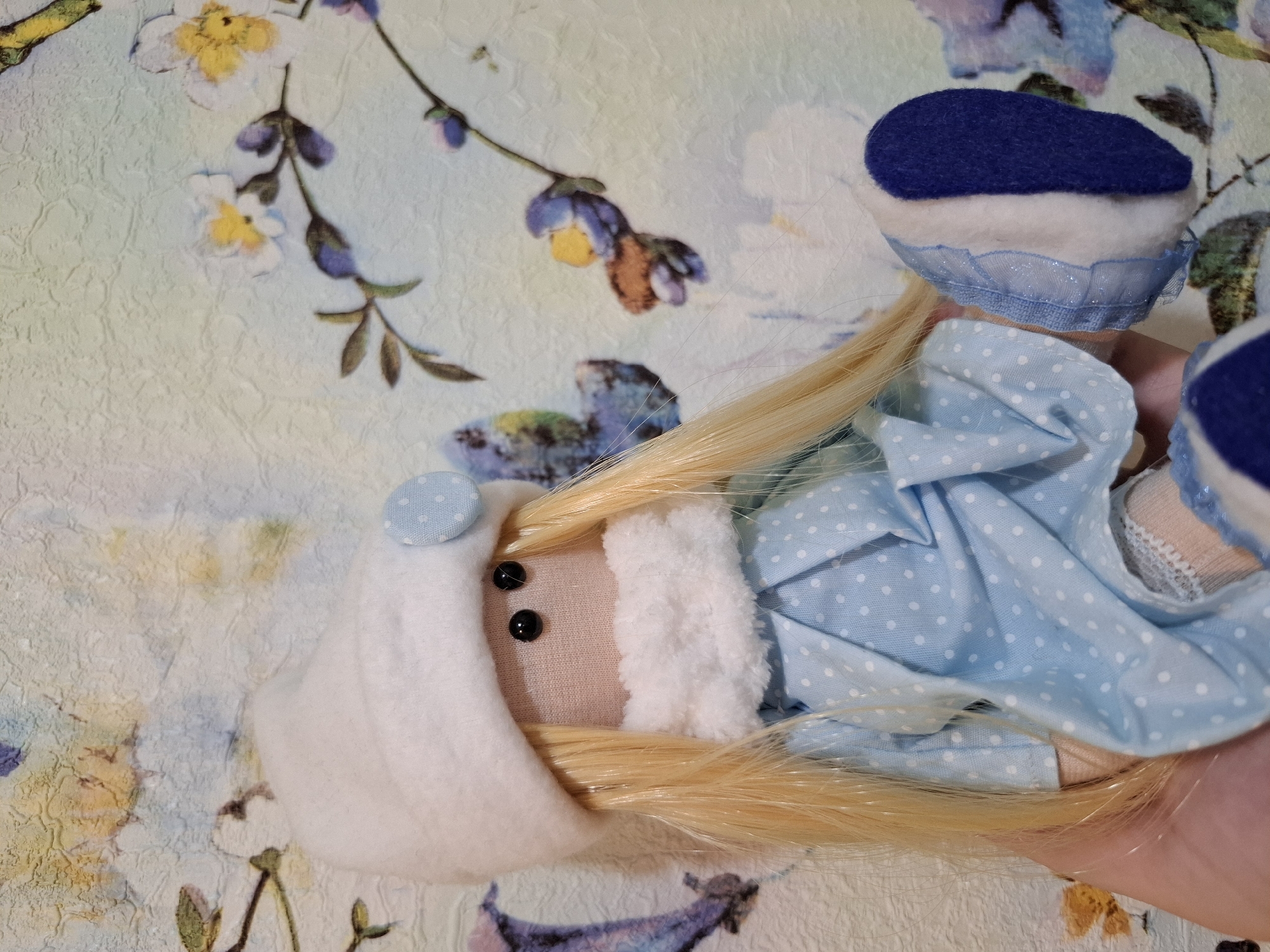 A close-up shows a handmade rag doll with a light complexion, blonde hair, and black button eyes lying on a floral patterned surface. The doll is wearing a white hood with a light blue polka-dotted button, a blue and white polka-dotted dress, and a blue beret with a white band. Part of a human hand can be seen in the lower right corner, presumably holding the doll. The setting appears to be indoors, with the patterned surface suggesting a decorative cloth or wallpaper. No information about the city of Амзя, Russia is present in the image.