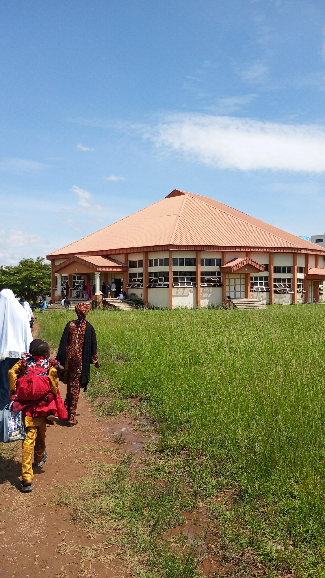 The image captures a sunny day in Jos, Nigeria, with a modern building featuring an orange, sloped roof dominating the background. The building's facade is a light color with orange trim, and it has numerous windows, some of which are open. Several people are seen entering and exiting the building, with some sitting on the steps. In the foreground, a path made of red dirt leads towards the building. To the left of the path, two individuals are walking away from the viewer. The person closest to the viewer is a child wearing a yellow outfit and a red backpack, carrying a blue bag. Behind the child, an adult in a dark headscarf and a colorful patterned dress walks. To the left of them, another person in a white headscarf and dark clothing is partially visible. The path is bordered on the right by tall, green grass that appears somewhat unkempt, with some puddles suggesting recent rainfall. A few trees are visible in the distance to the left of the building. The sky is a clear blue with a few white clouds.