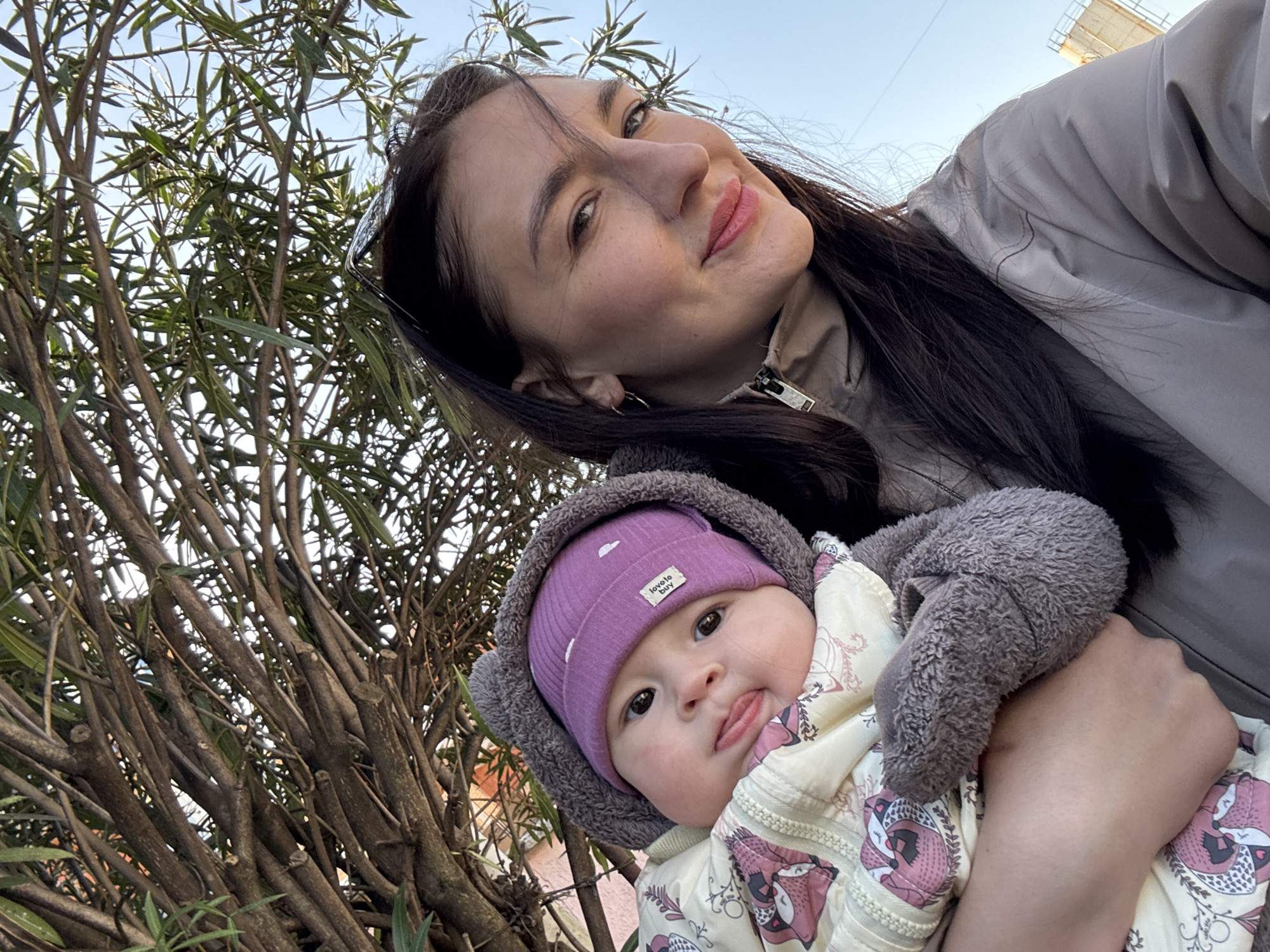 A medium-shot selfie features a woman holding a baby. The woman is light-skinned with dark hair. She is wearing a light-colored jacket and has sunglasses pushed up on her head. The baby, held in her arms, is wearing a purple beanie and a patterned, puffy jacket with gray furry trim. The baby is sticking its tongue out. Behind them is a dense, multi-stemmed bush. The sky is visible in the background. Based on visual characteristics and common elements, this photo appears to have been taken in Sochi, Russia.