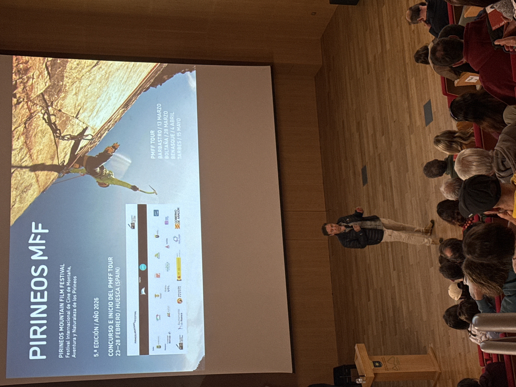 A medium shot depicts a presentation about the Pirineos Mountain Film Festival (MFF) in front of an audience. On the left side of the frame, a large screen displays the festival's logo and information, including that it is the 5th edition in the year 2026. The screen also advertises the PMFF Tour with dates for Barbastro, Boltaña, Benasque, and Tarbes. A man stands to the right of the screen, facing the audience and gesturing with his hands as he speaks. Seated in rows, the audience faces the presentation. The room has wooden floors and walls, with a simple wooden podium in the lower-left corner. The location is identified on the screen as Huesca, Spain, but Benasque is not explicitly mentioned in the image.