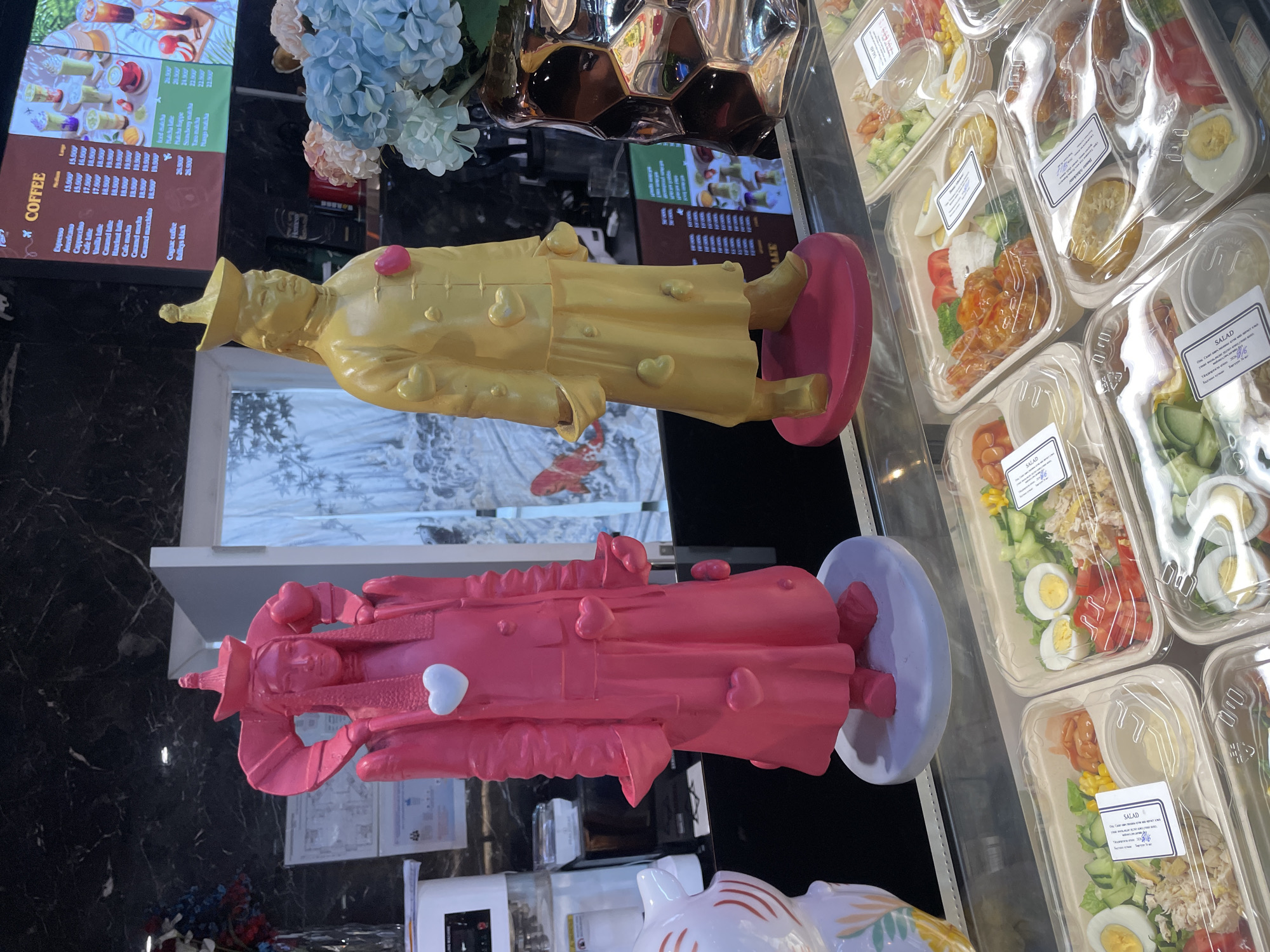 In Sergelen, Mongolia, a brightly lit display case showcases an assortment of pre-packaged salads. Behind the salads, two statues stand prominently. The statue on the left is a figure in a vibrant pink robe, adorned with heart shapes of various sizes. The statue on the right is a figure in a golden robe, also decorated with heart shapes, and wearing a traditional hat. A large menu board with "COFFEE" clearly visible hangs on the wall behind the statues, displaying a variety of coffee and matcha-based beverages with prices in a foreign currency. The environment appears to be a food establishment, possibly a cafe or restaurant, with a polished dark countertop and tiled wall.