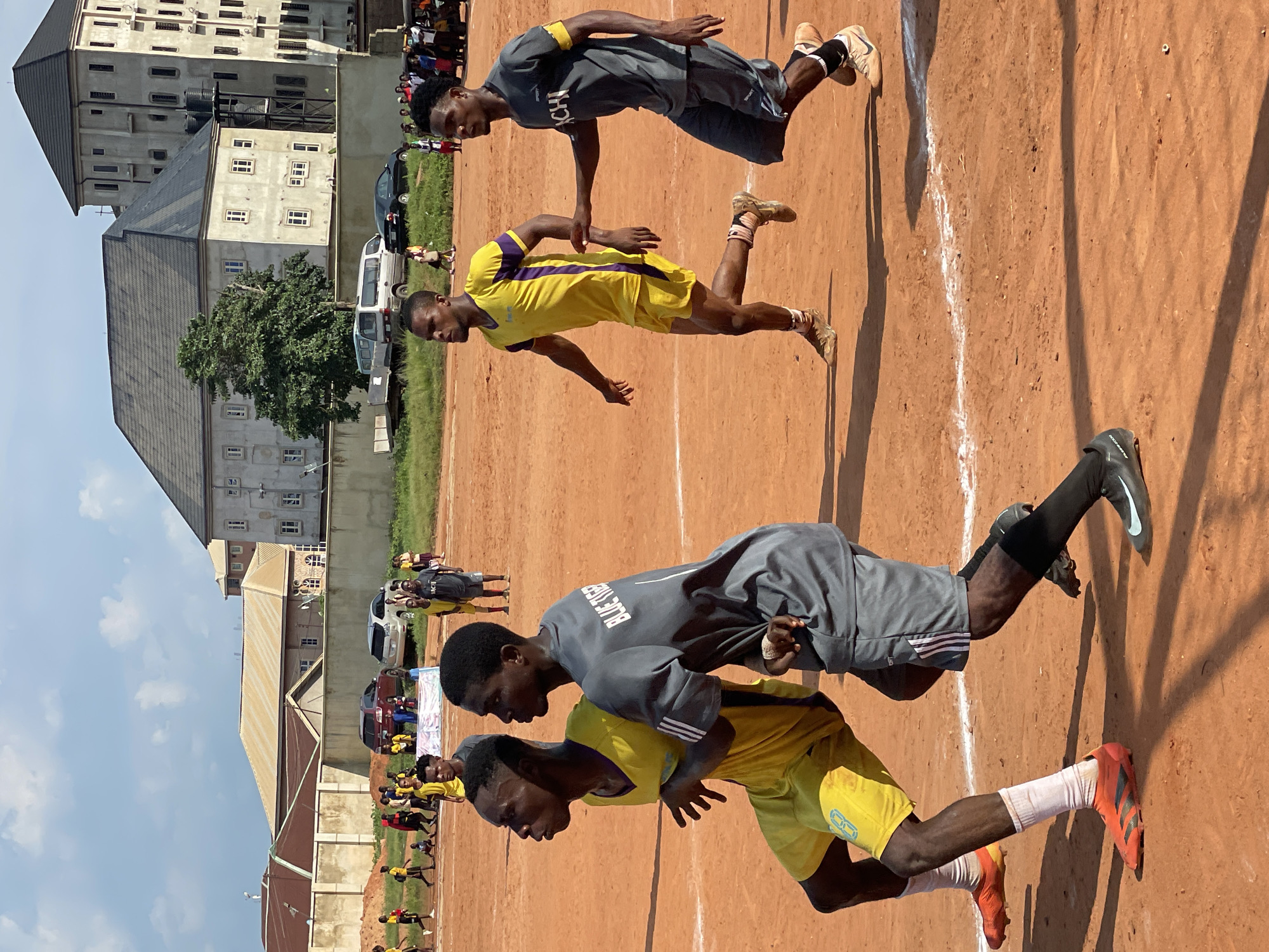 An outdoor sports field with a clay surface is the setting for this image. In the foreground, two male athletes are running. The athlete on the left is wearing a yellow jersey with purple accents and yellow shorts, and the athlete on the right is wearing a gray jersey with white lettering that says "BLUE TIGER" and gray shorts. Both are wearing athletic shoes. In the midground and background, other individuals, some in similar athletic attire, are present on the field. Buildings, including a multi-story structure and a longer, lower building with a tan roof, are visible behind a wall in the background. Vehicles, including cars and a van, are parked near the buildings. The sky is partly cloudy.